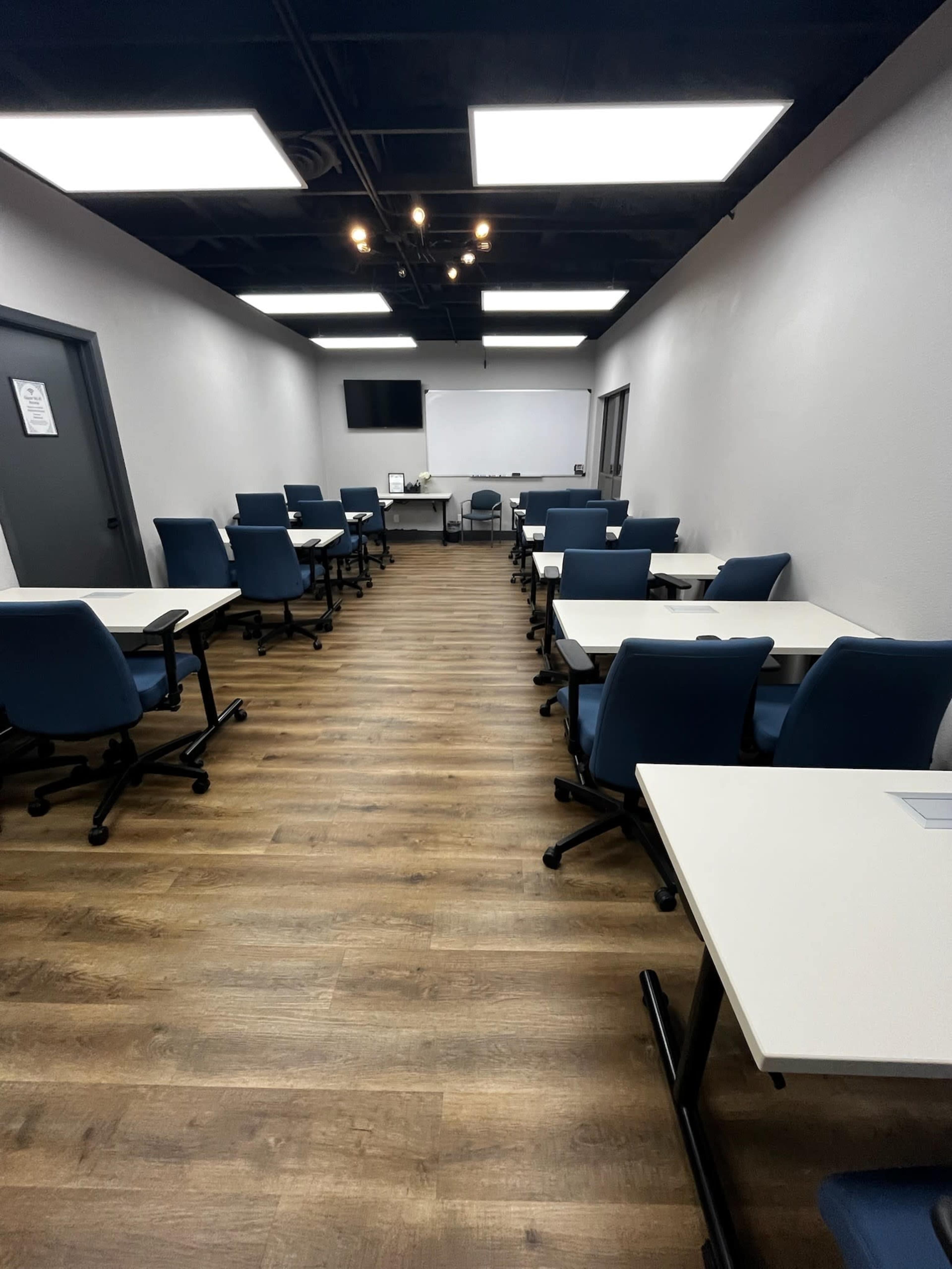 The image shows a classroom or meeting room with several rows of tables and chairs, all arranged neatly, and equipped with a large screen and whiteboard at one end.