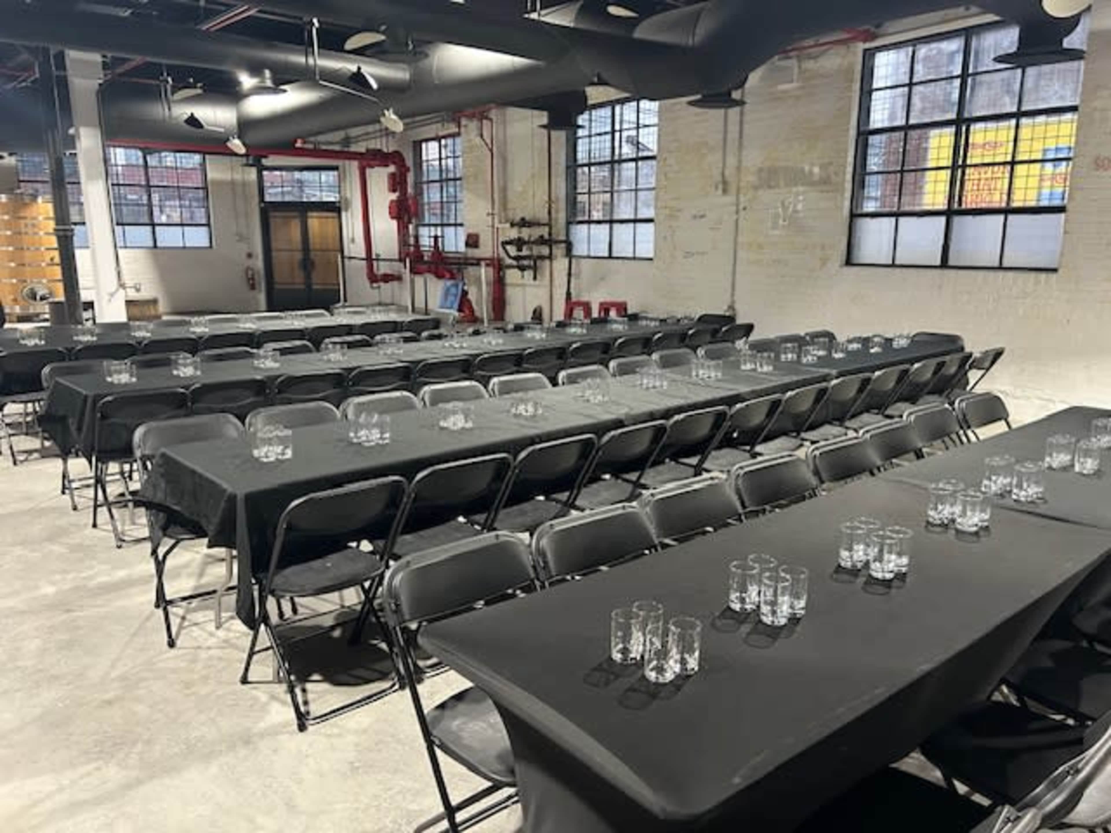 A large event space with multiple rows of black tables set with glassware, arranged in a grid layout.
