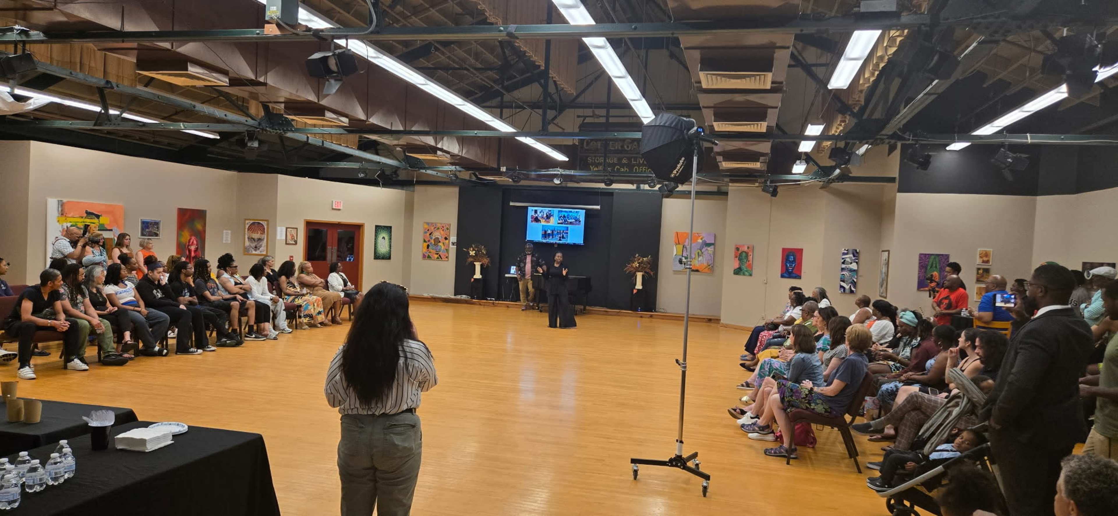 A diverse group of people gathers in a large room with a wooden floor, facing a stage where speakers present to the audience.