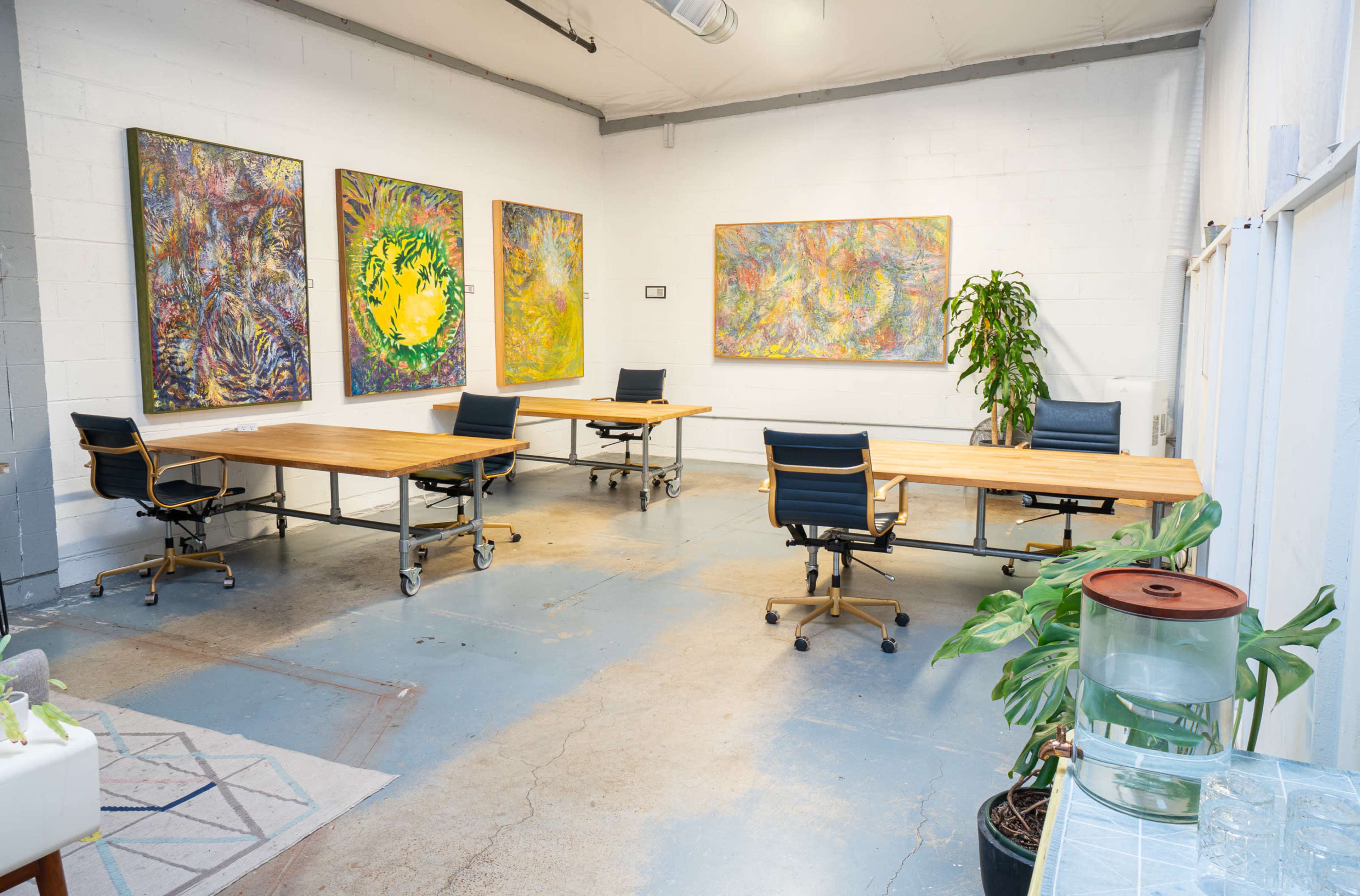 The image shows a spacious office area with four wooden desks, rolling chairs, colorful artwork on the walls, and a large plant in the corner.