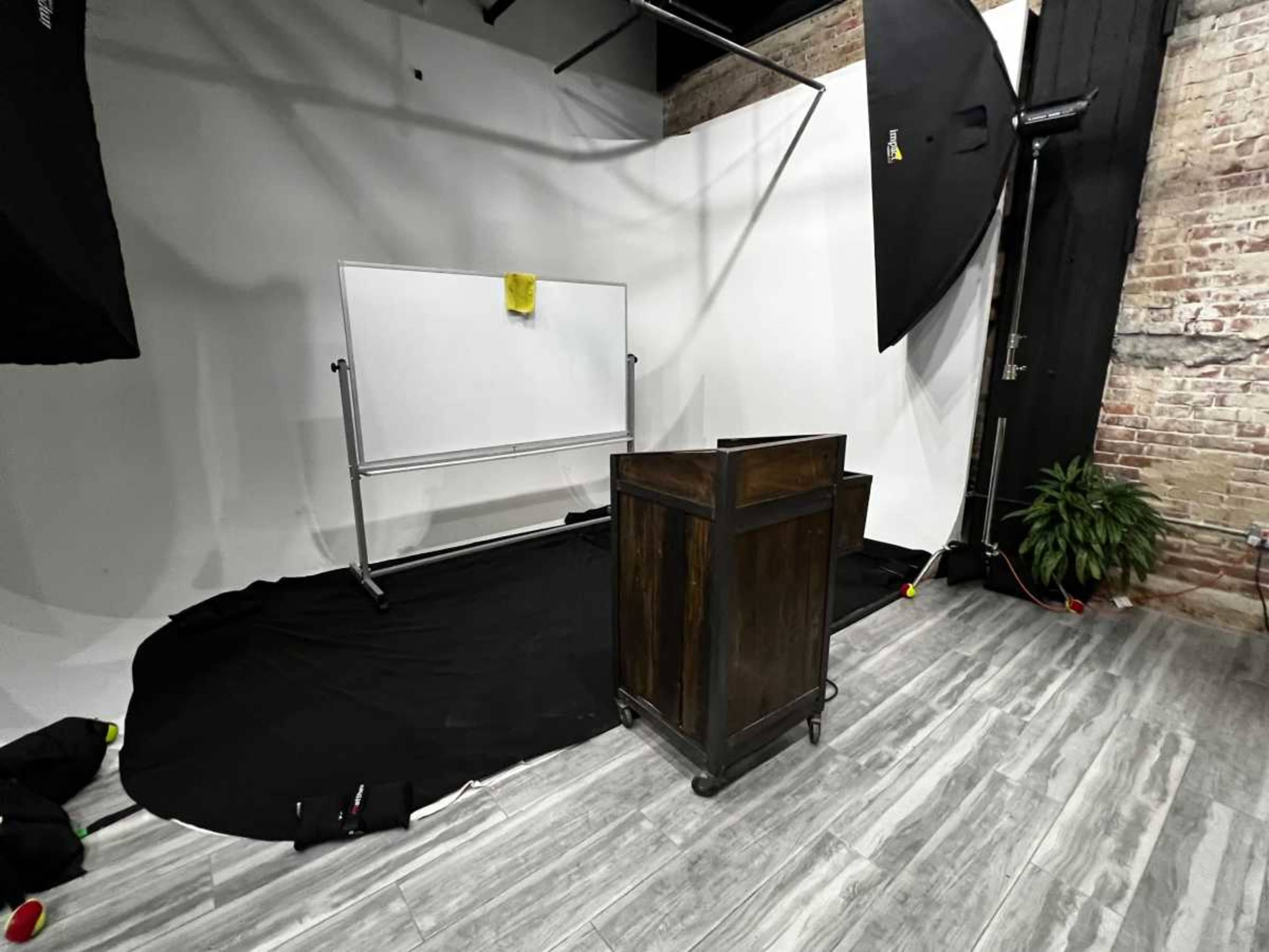 The image shows a studio setup featuring a wooden podium facing a blank whiteboard against a backdrop of light-colored walls and a floor covered with black fabric.