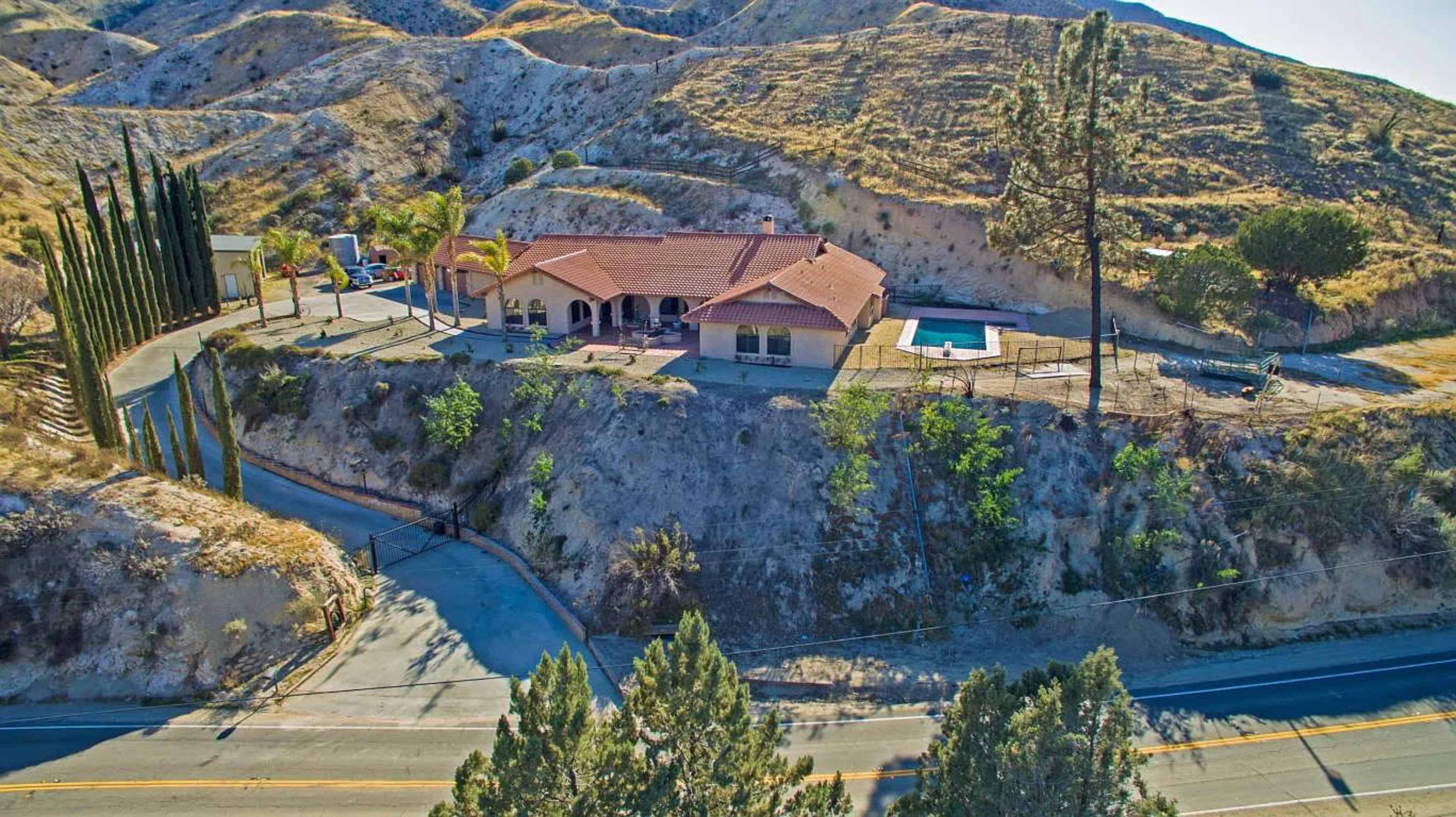 A single-story house on a hillside with a swimming pool, surrounded by trees and a winding road below.