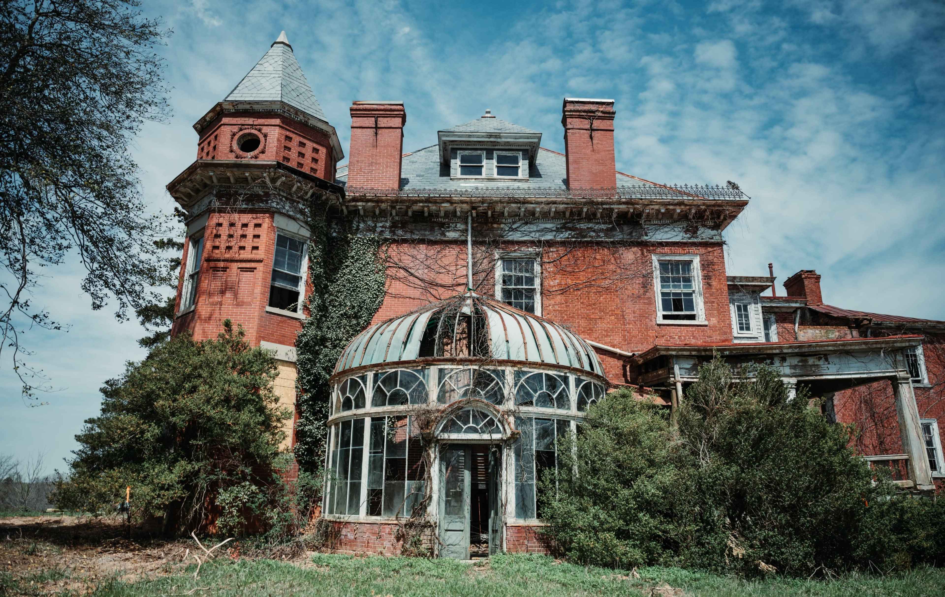 The image shows a large, dilapidated brick mansion with a glass conservatory, surrounded by overgrown vegetation under a cloudy sky.