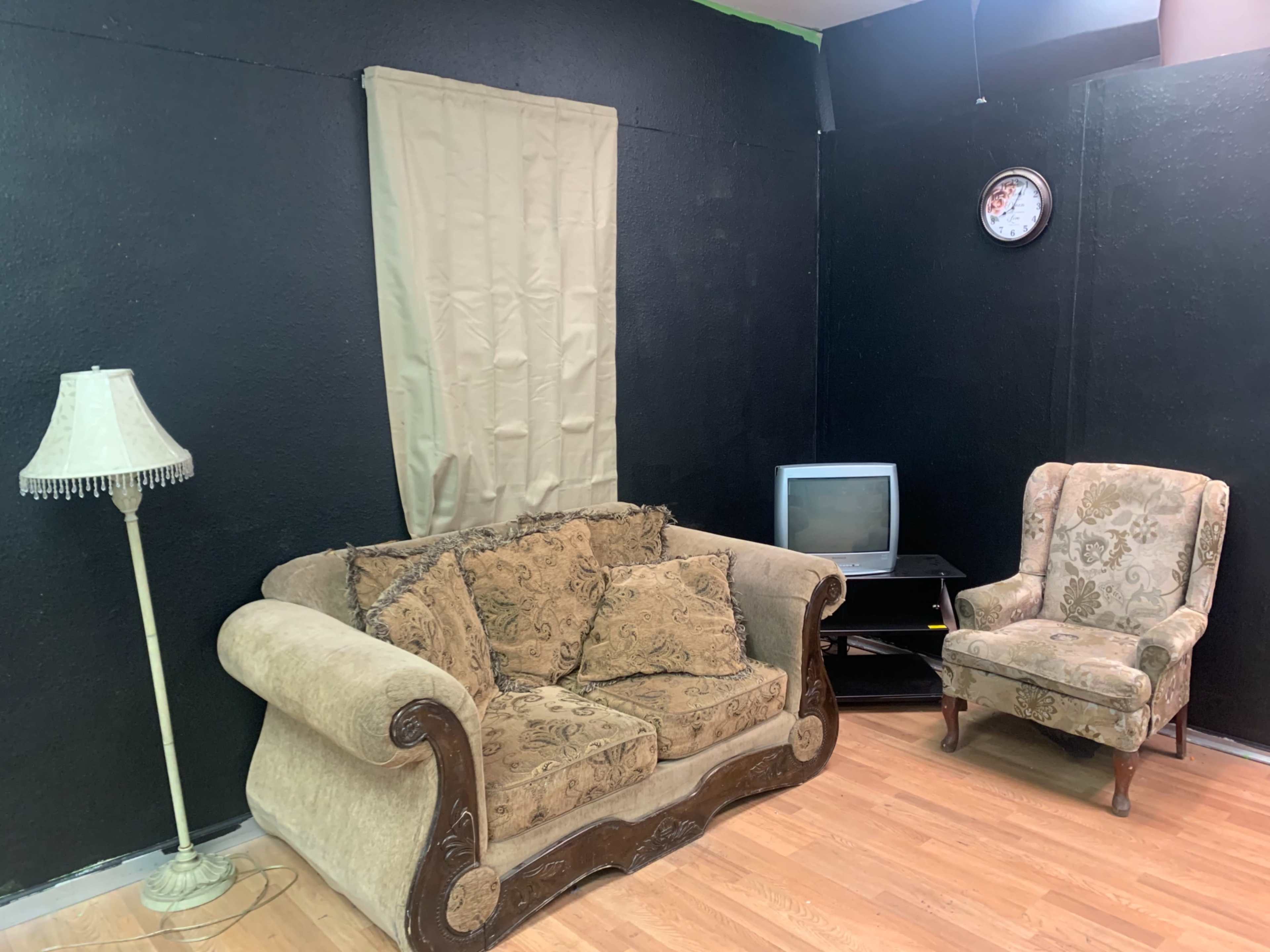 A small living area with a lamp, a patterned armchair, a vintage-style couch with cushions, a television on a stand, and a clock on the wall, all set against a dark wall with a curtain.