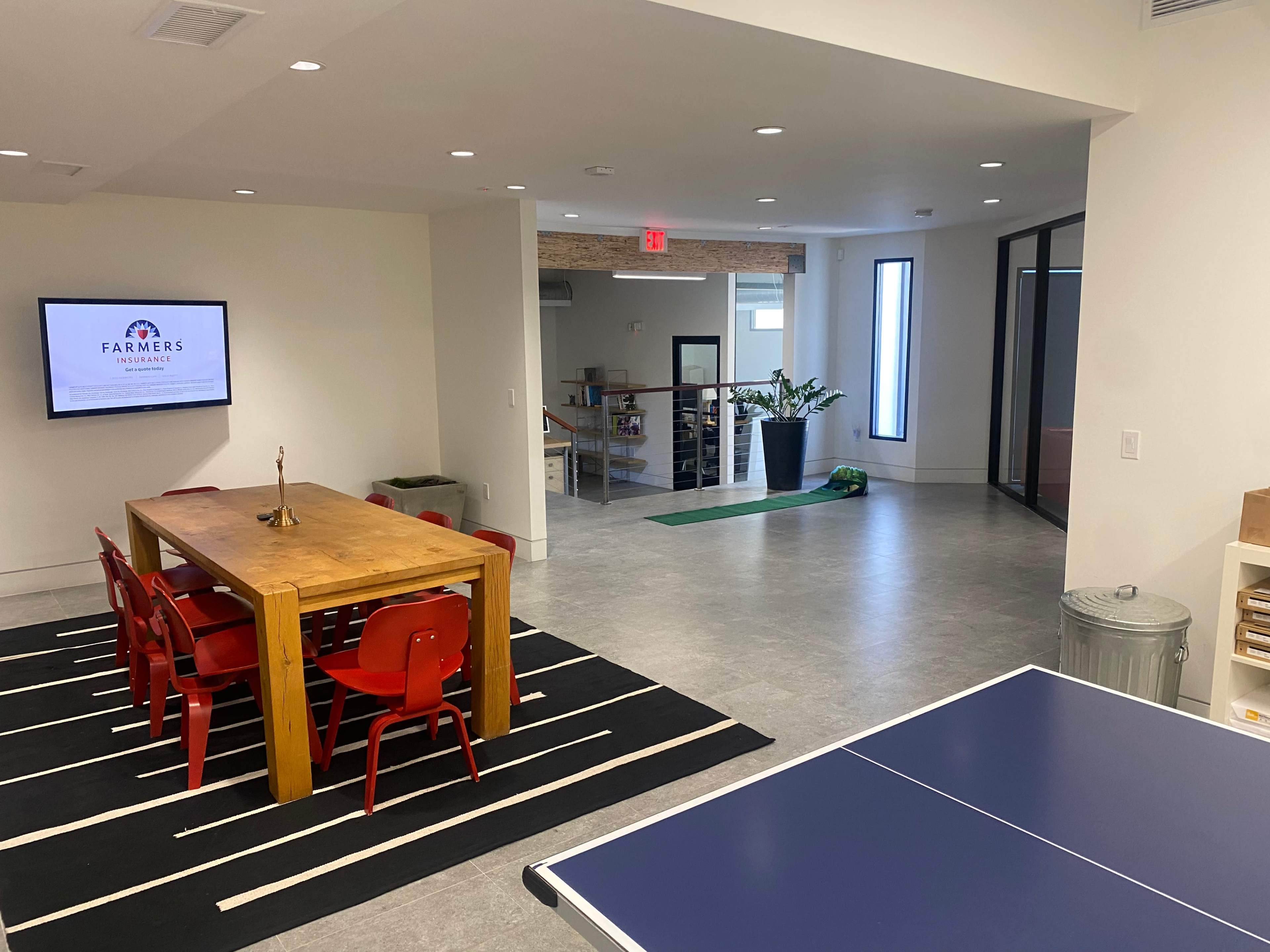 A modern interior space featuring a wooden dining table with red chairs, a ping pong table, and a large television displaying a logo, with plants and windows in the background.