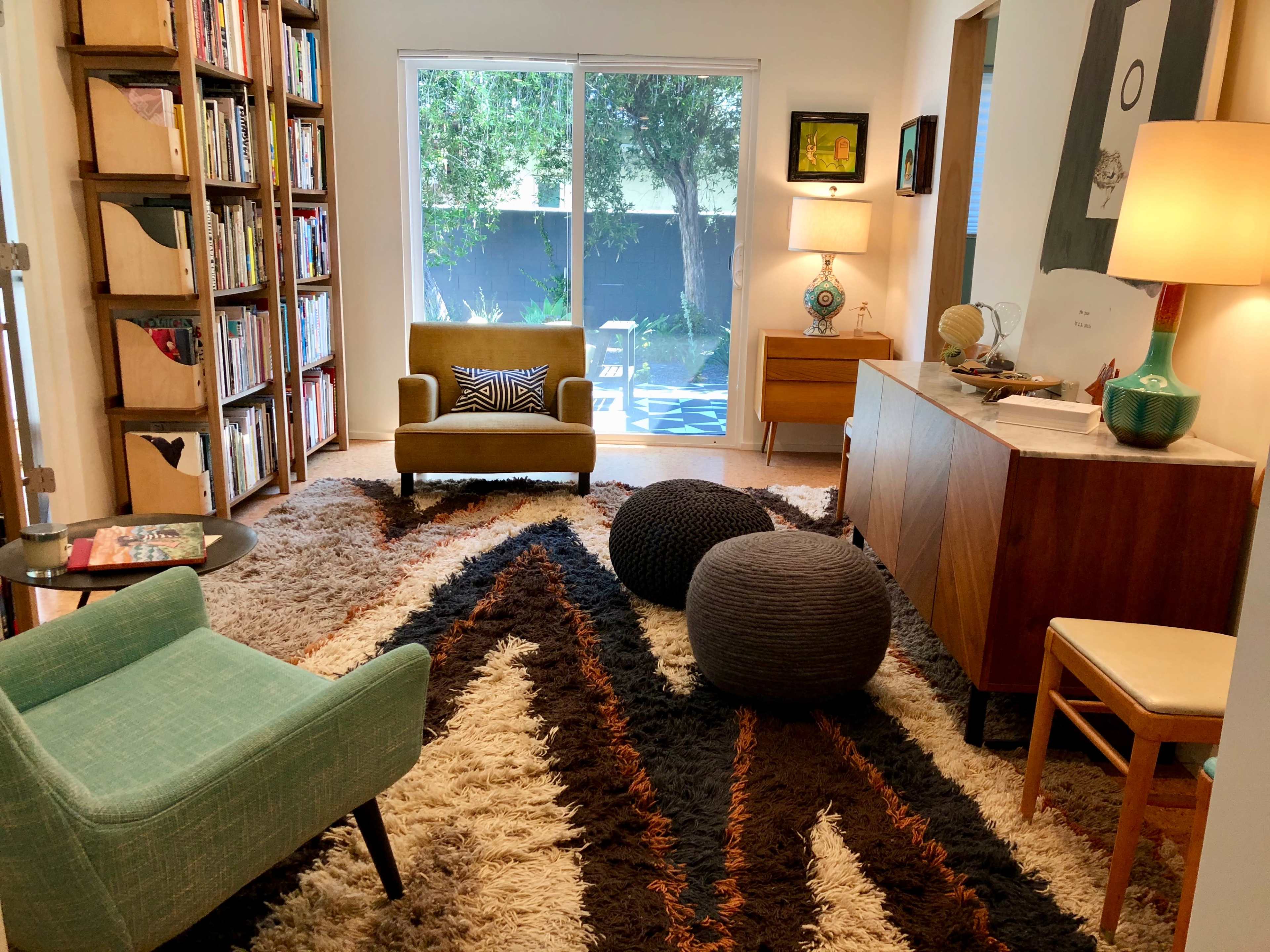The image shows a cozy living room with a bookshelf, armchair, and a patterned rug, featuring a large window that opens to a garden view.