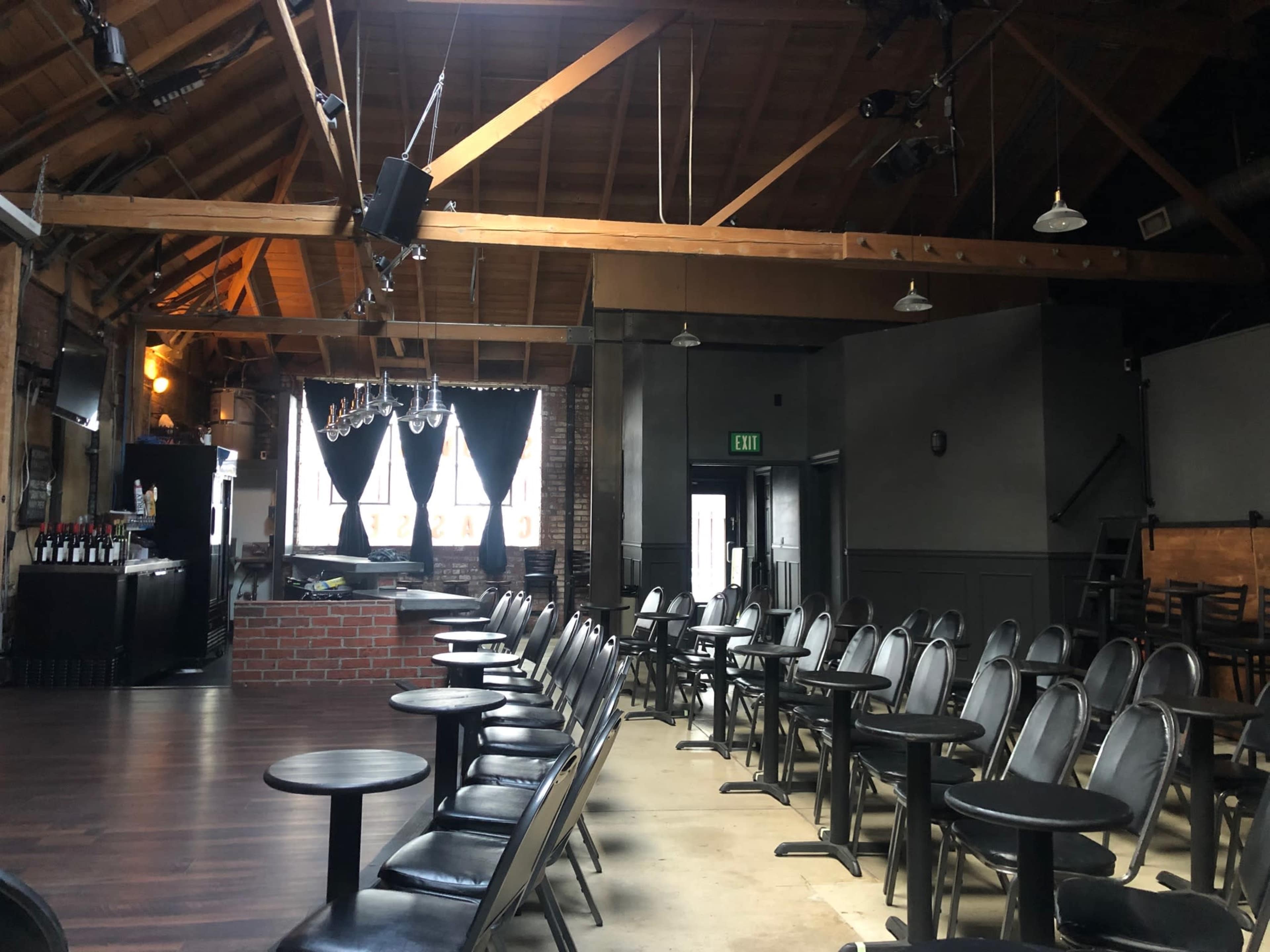 A spacious venue with black chairs and small tables arranged in front of a bar area beneath a wooden ceiling.