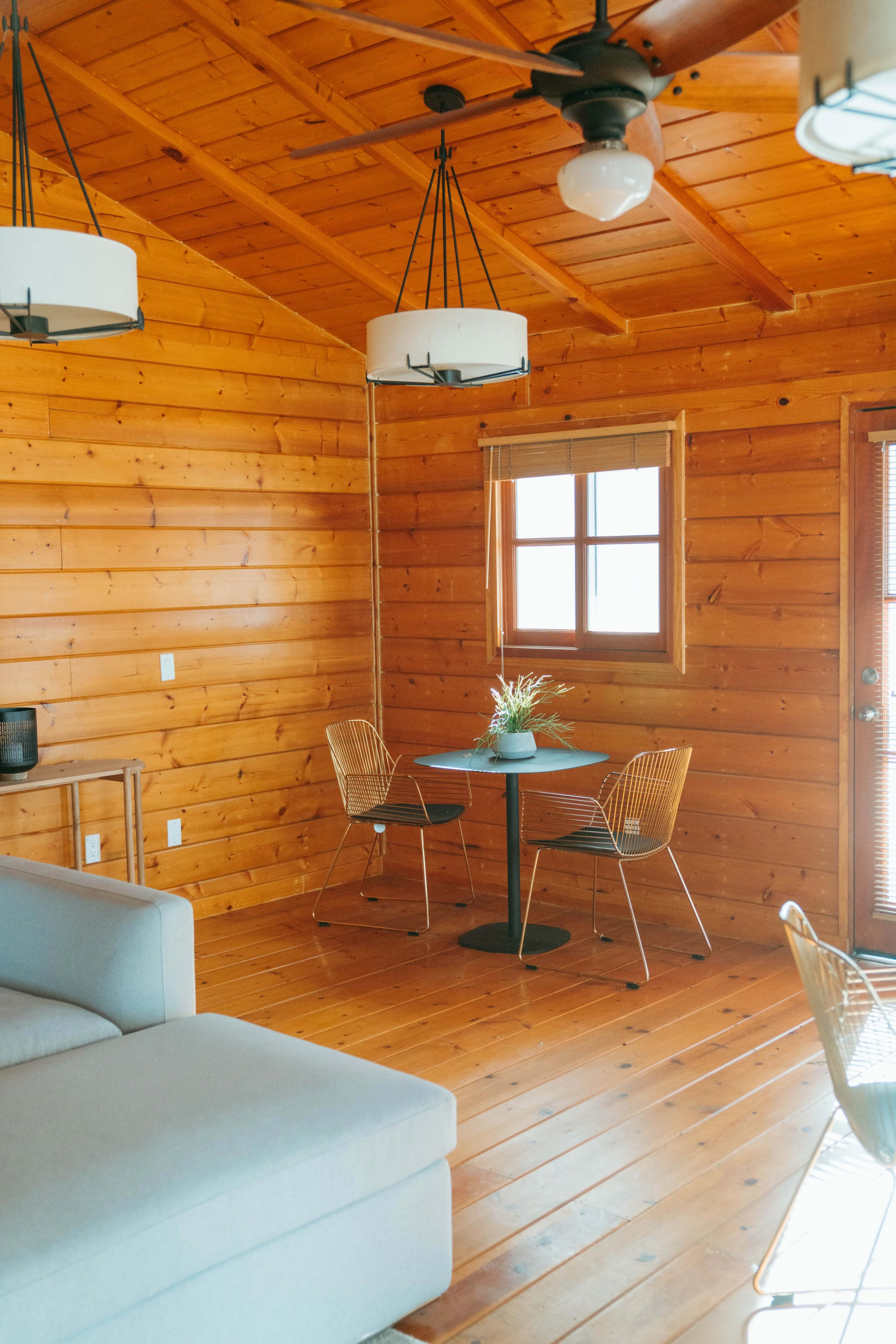 A wooden interior of a cabin with a small dining table and two chairs, a sofa, and large pendant lights hanging from the ceiling.