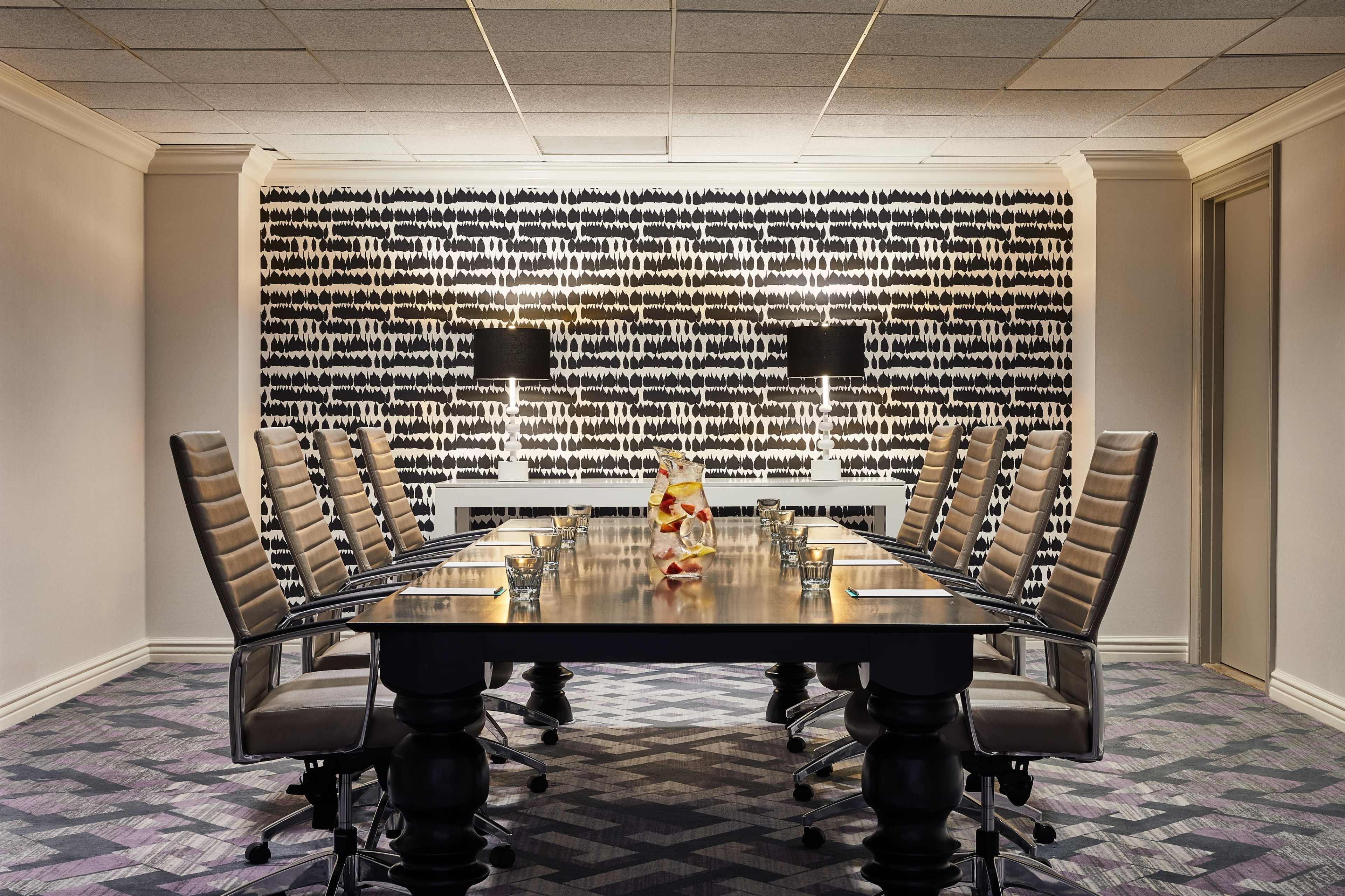 A modern conference room features a long table surrounded by high-backed chairs, with a patterned wall and two lamps providing ambient light.