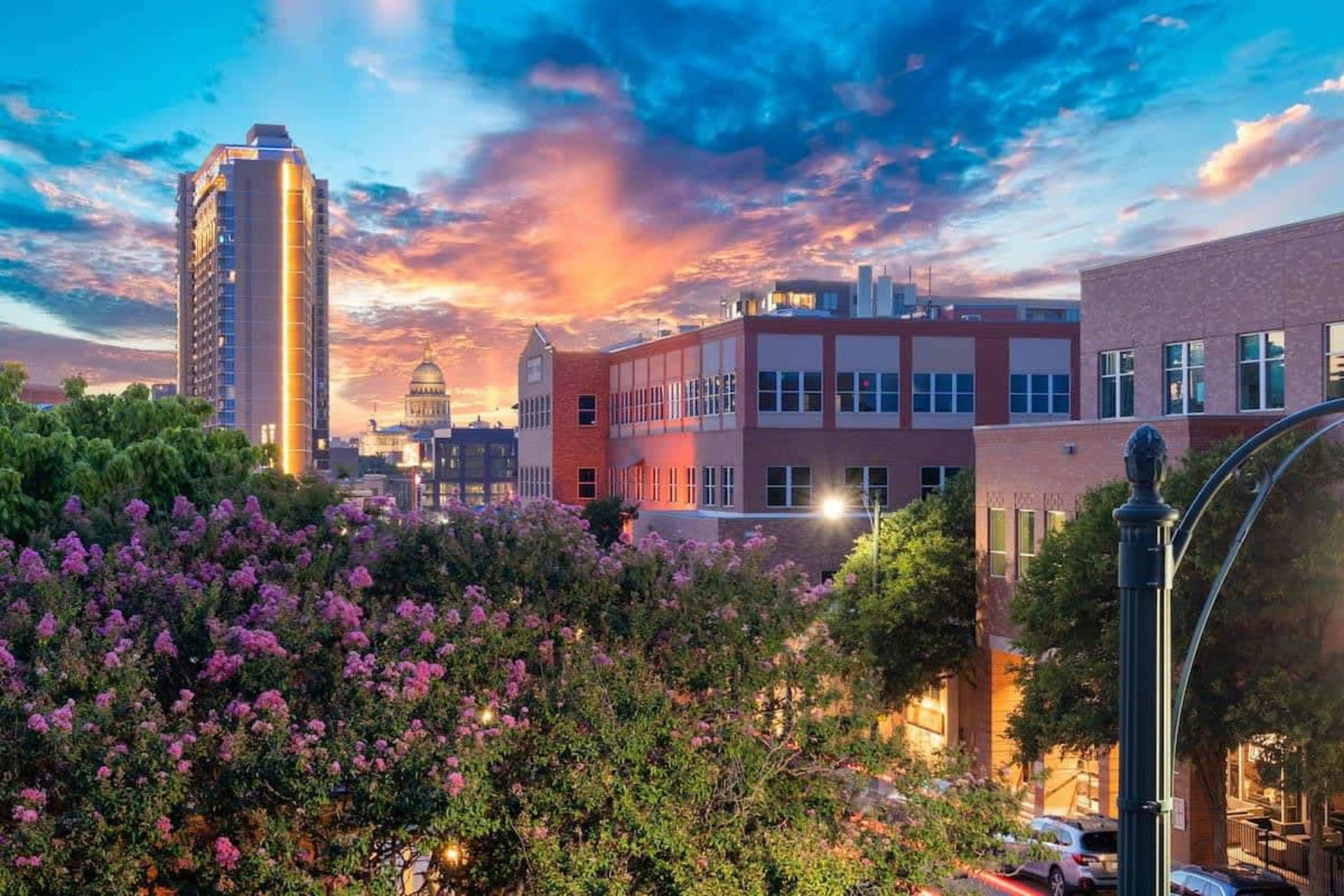 The image shows a cityscape during sunset with buildings, trees, and vibrant clouds in the sky.
