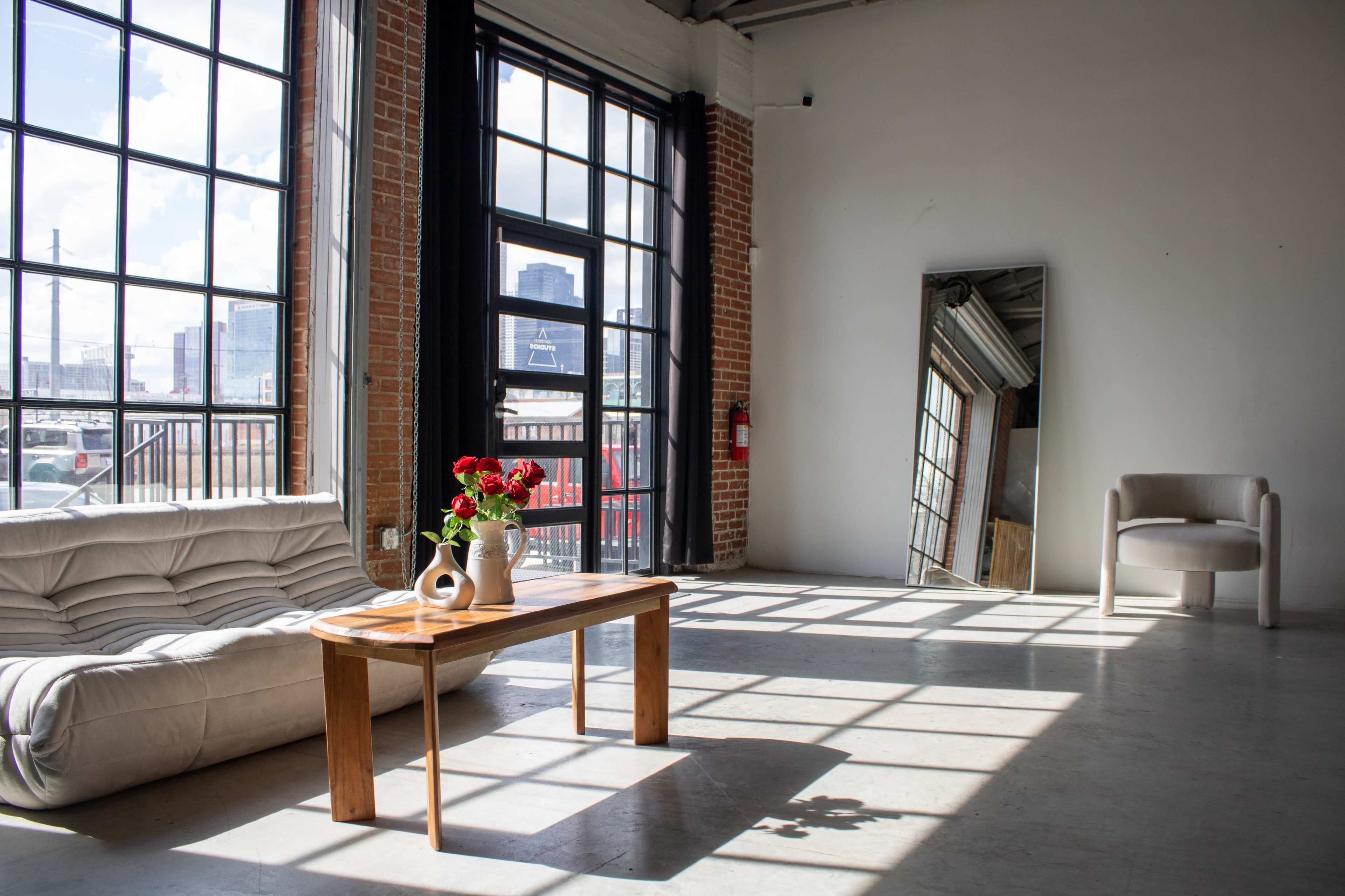 The image shows a bright, spacious living area featuring a couch, a wooden coffee table with a vase of flowers, a large mirror, and a light-colored chair next to large windows.