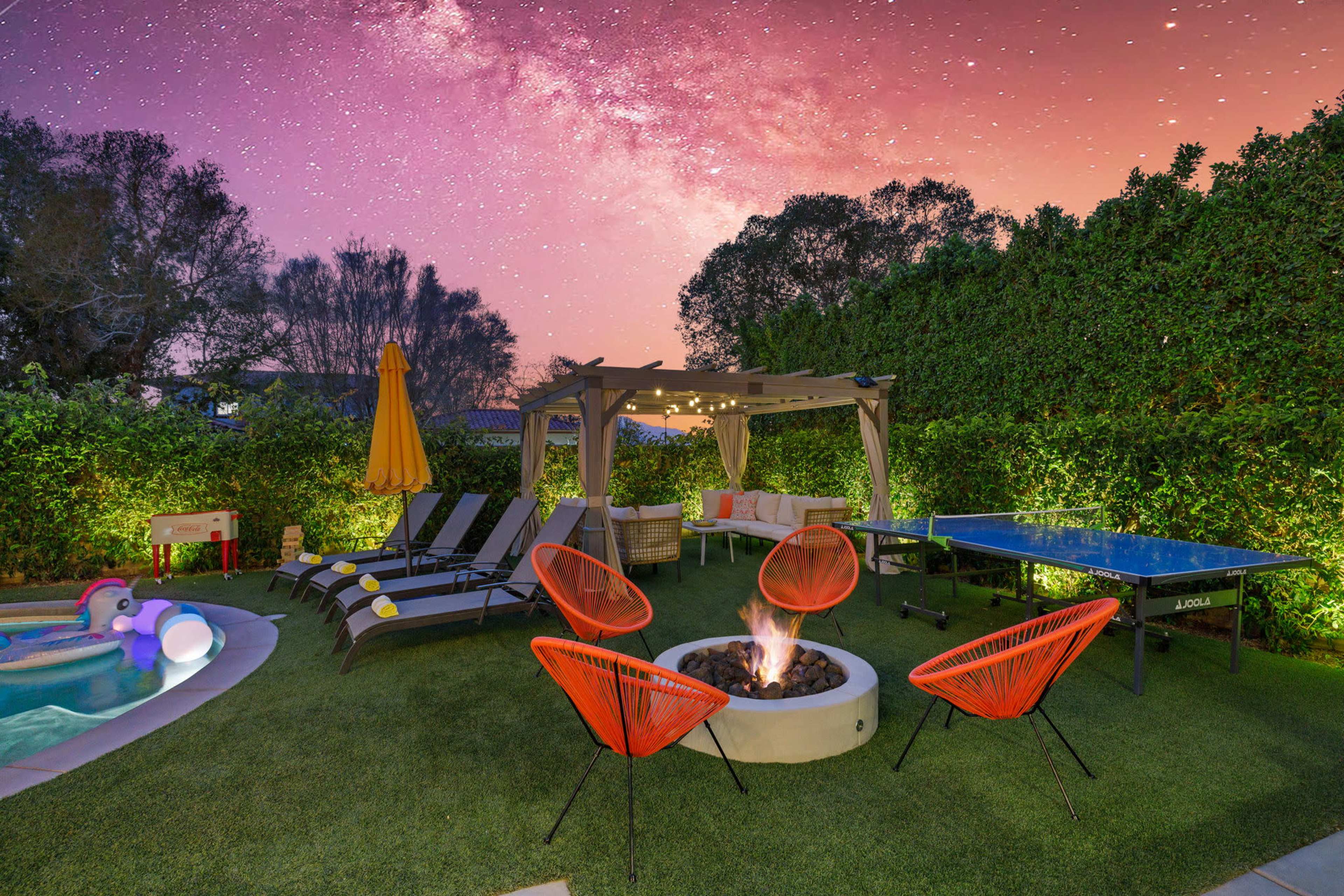 A backyard features a fire pit surrounded by chairs, a swimming pool, a ping pong table, and outdoor seating under a pergola, set against a starry sky.