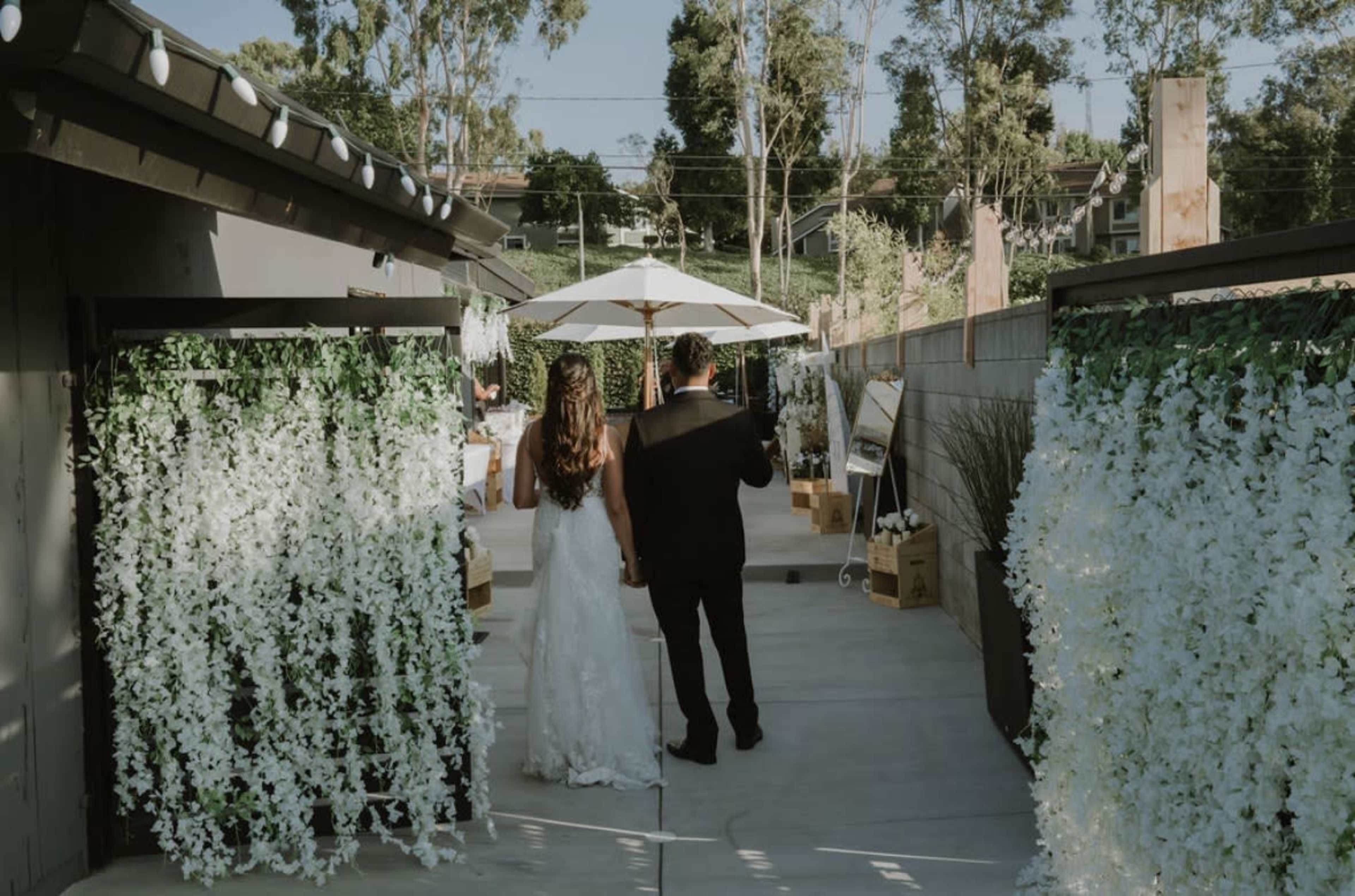 A bride and groom walk hand in hand down a pathway lined with white floral decorations and greenery, under a sunshade umbrella.