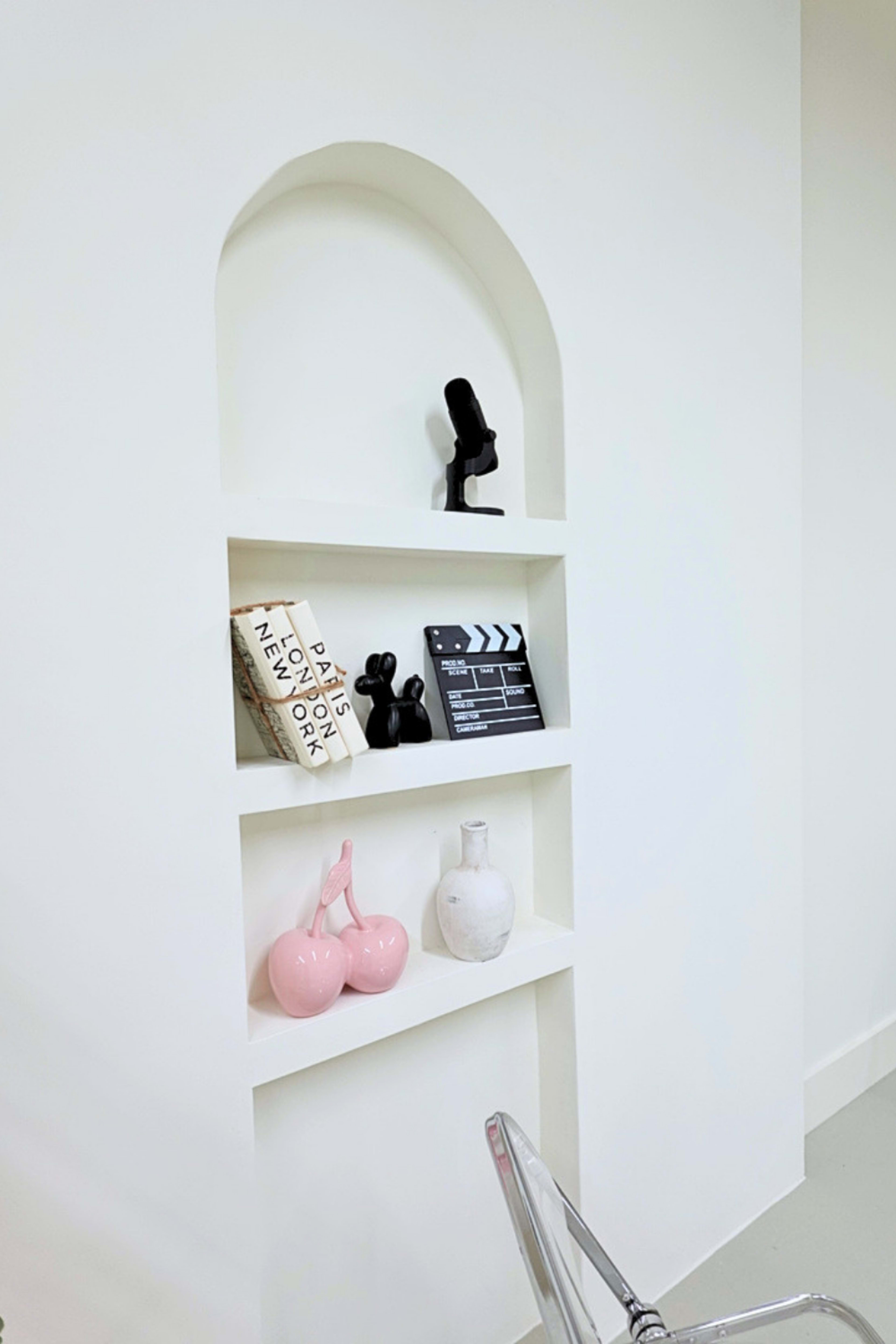 The image shows a white wall niche with three shelves displaying a book, a film clapperboard, a microphone, and decorative items, including two pink objects.