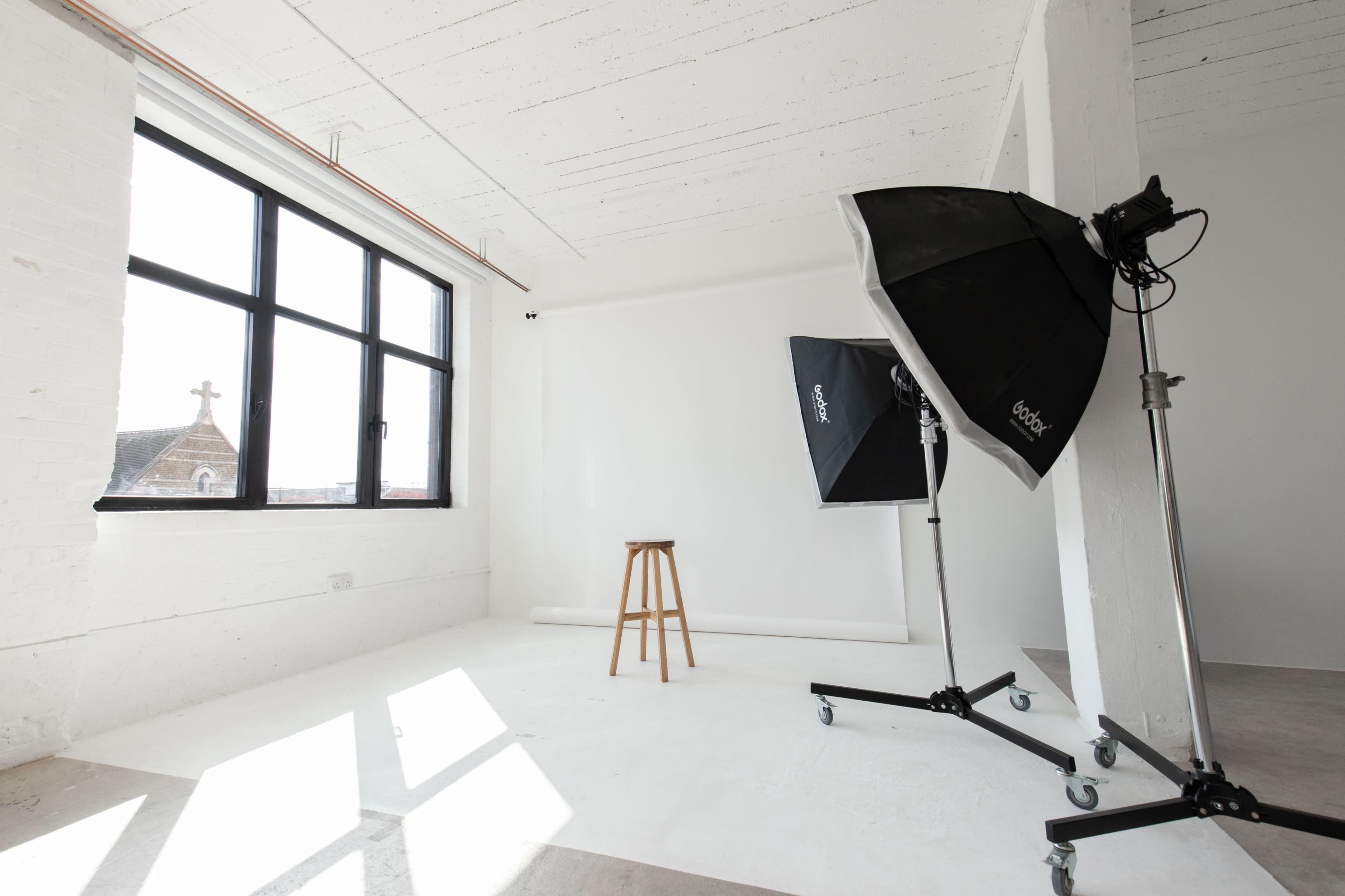 A photography studio with a wooden stool, two lighting umbrellas, and large windows allowing natural light.
