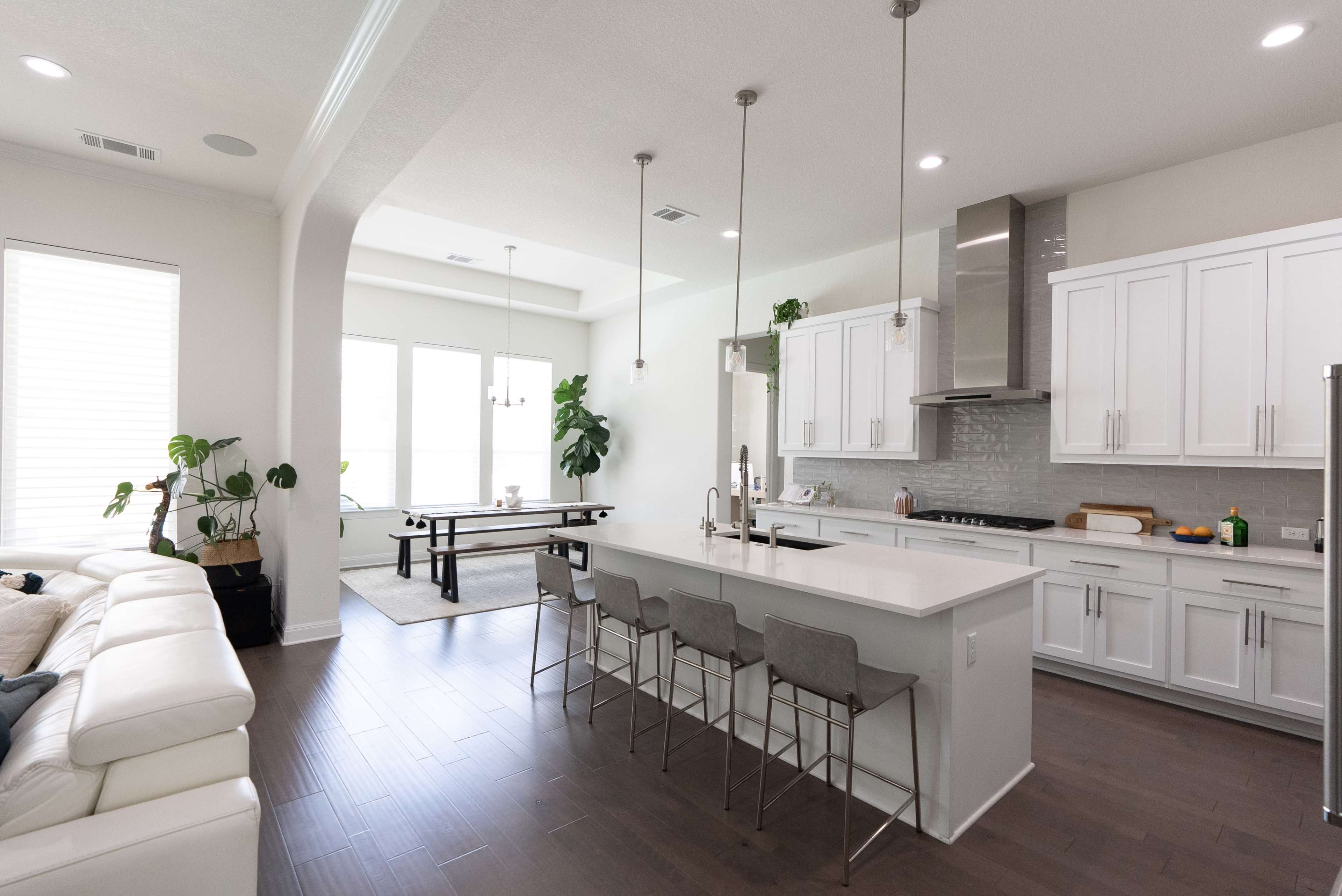 The image shows a modern kitchen with white cabinetry, stainless steel appliances, and a spacious dining area featuring a wooden table and a comfortable sofa nearby.