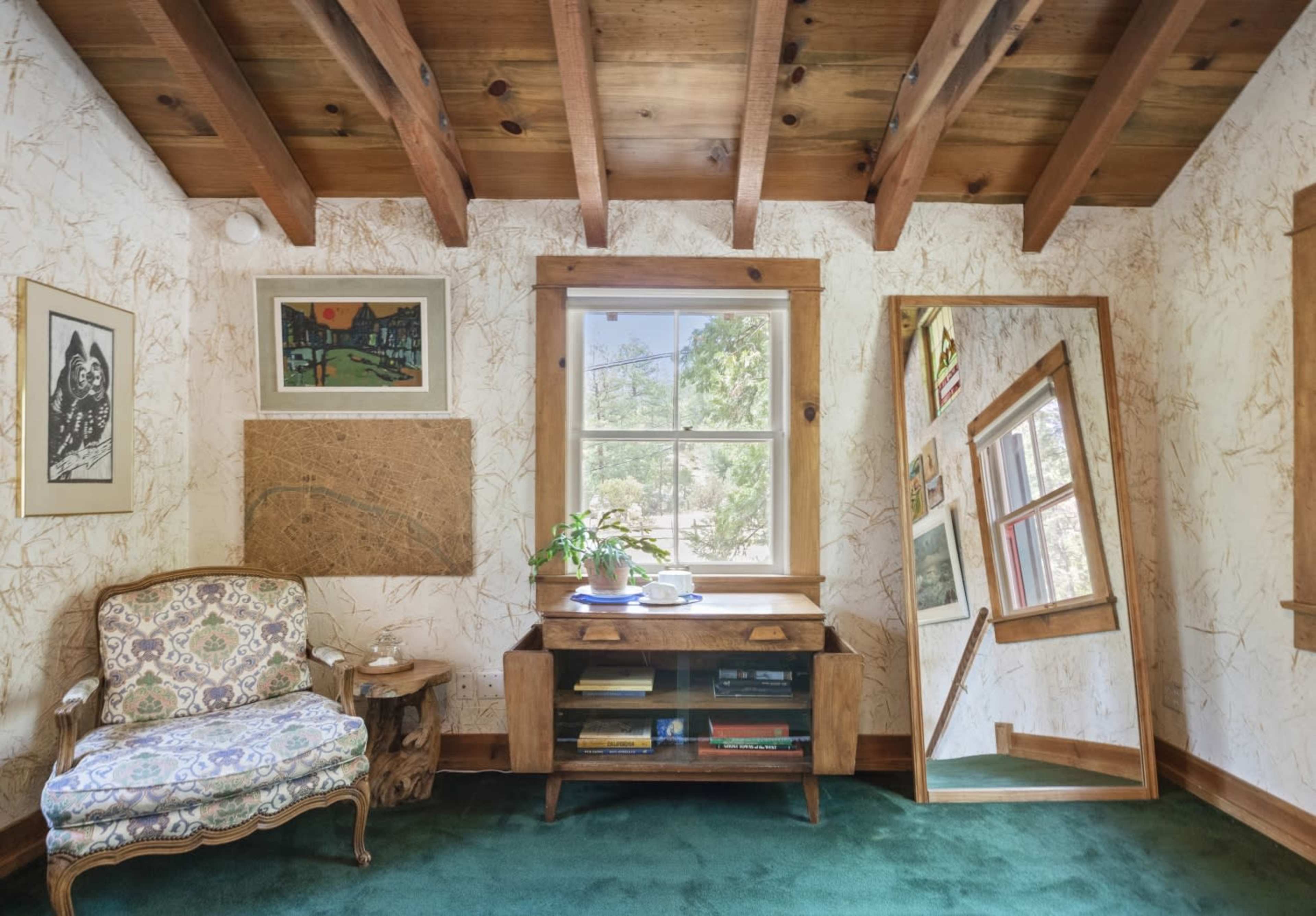 The image shows a room with wooden beams, a patterned chair, a wooden desk with books, and a large mirror beside a window.
