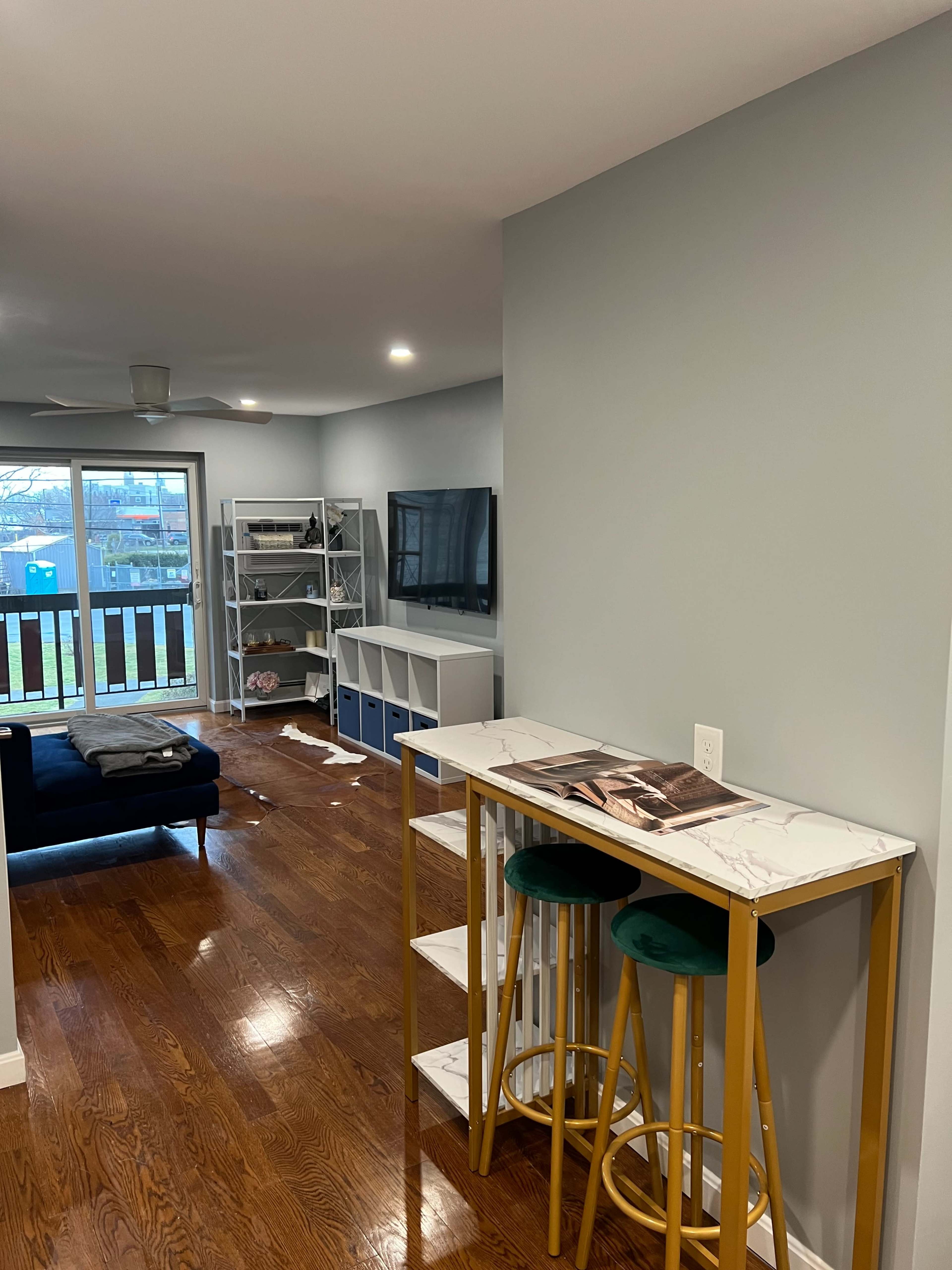 A modern living area featuring a small bar table with two stools, a dark blue sofa, a mounted TV, and shelves along the walls.