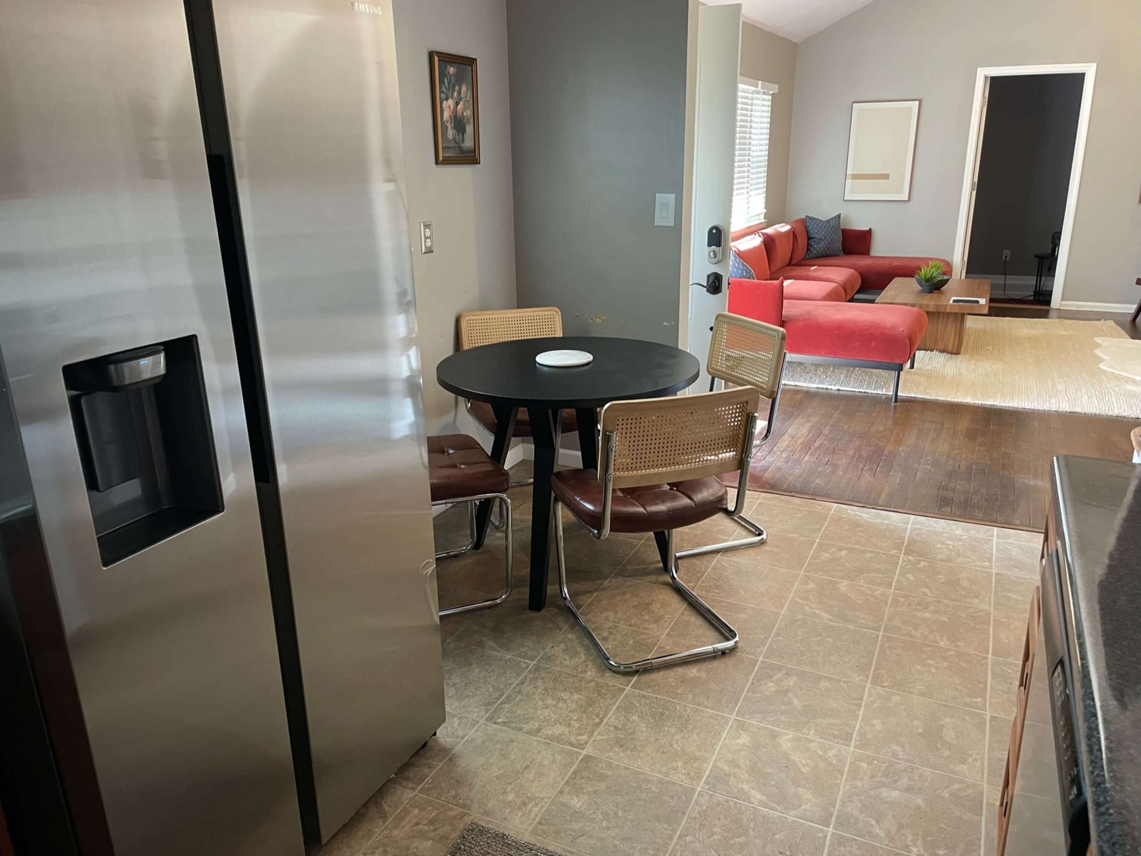 A kitchen with a round black table and four chairs is adjacent to a living room featuring a red sofa and a partially visible artwork on the wall.