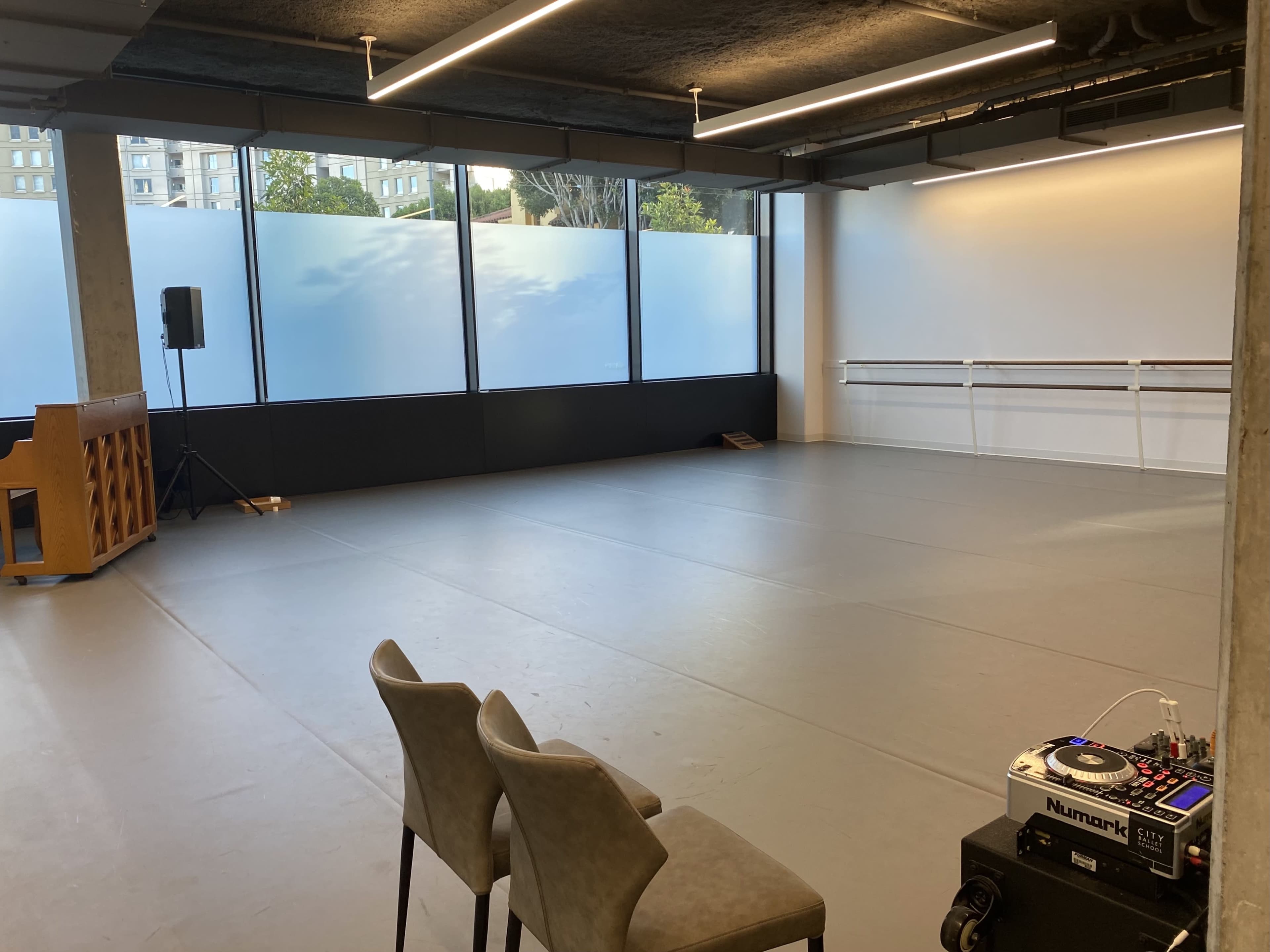 An empty dance studio with large windows, a wooden piano, two chairs, and a DJ controller set up on the floor.