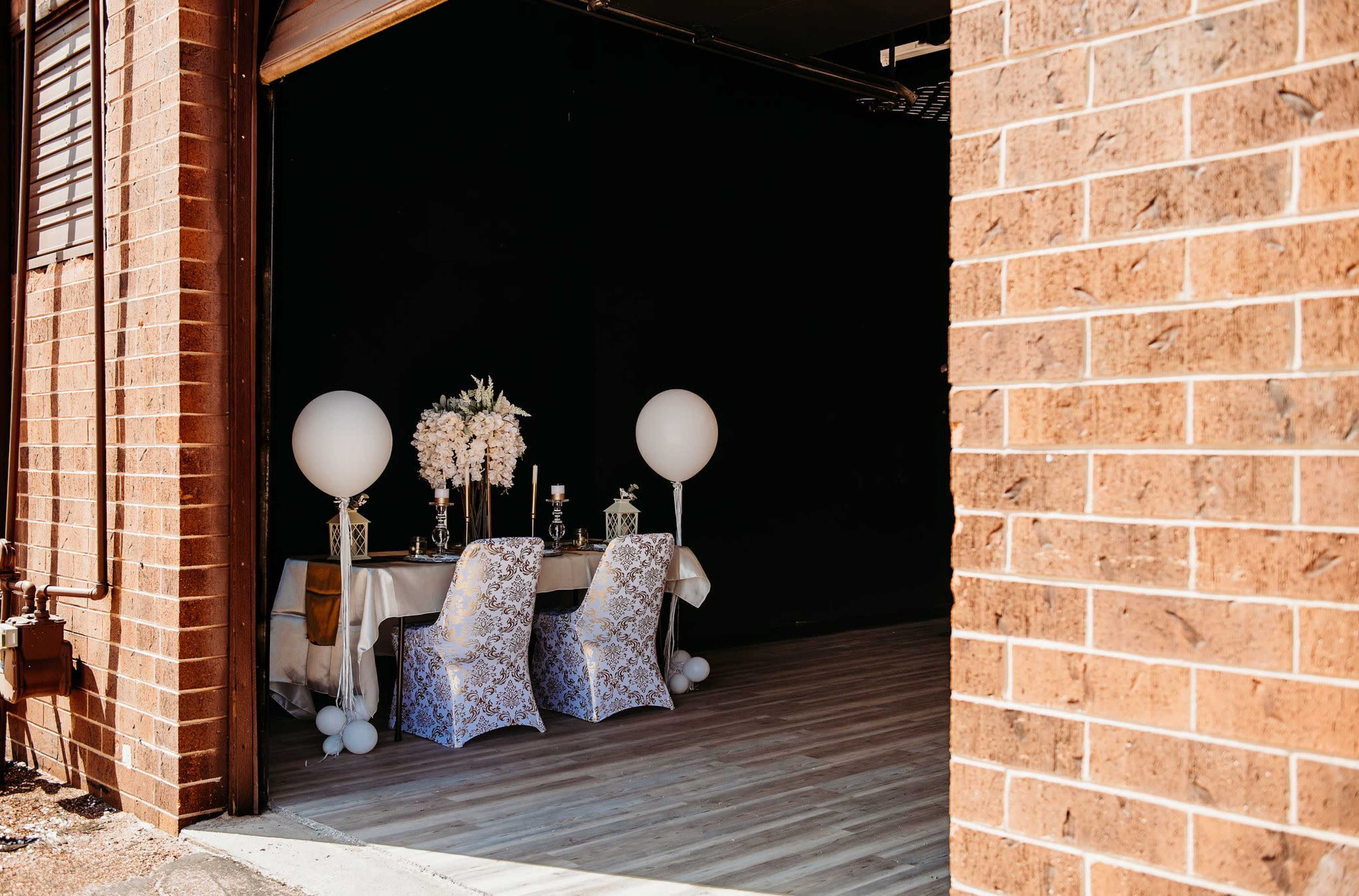 The image shows a decorated table set up with elegant chairs and large white balloons, visible through an open garage door against a dark background.