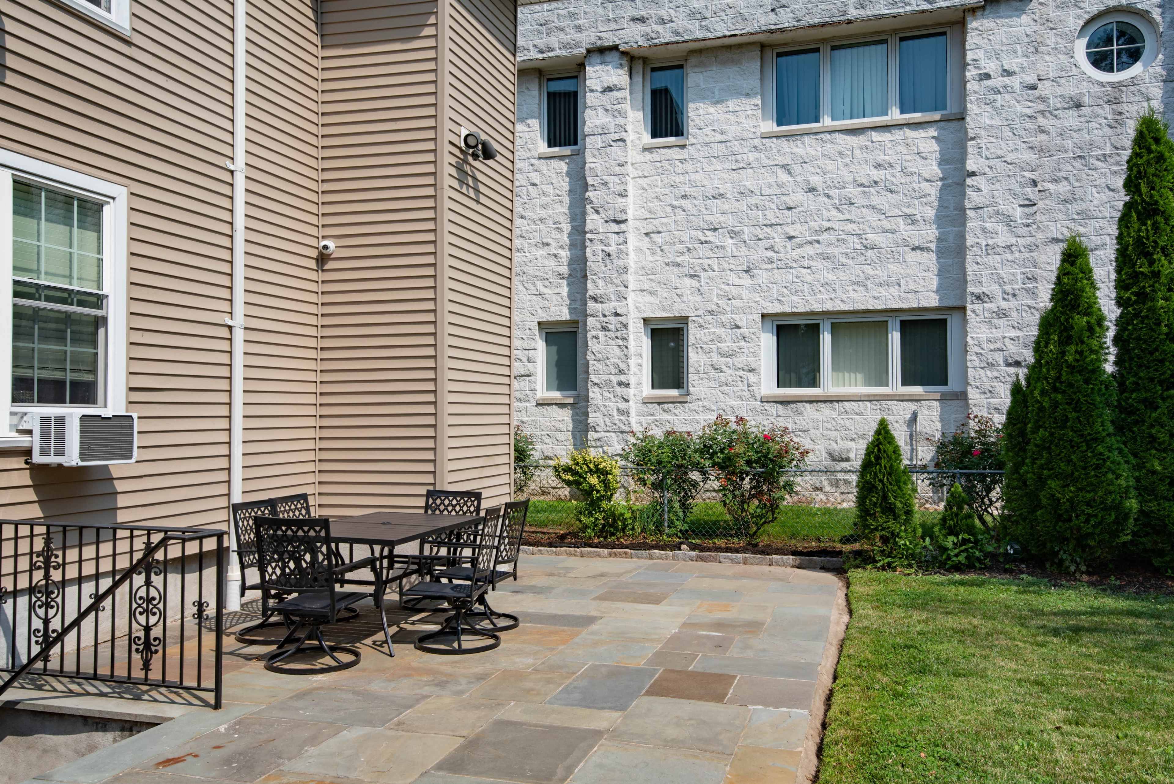 The image shows a stone patio with a metal table and chairs adjacent to a beige building and a white concrete structure in the background.