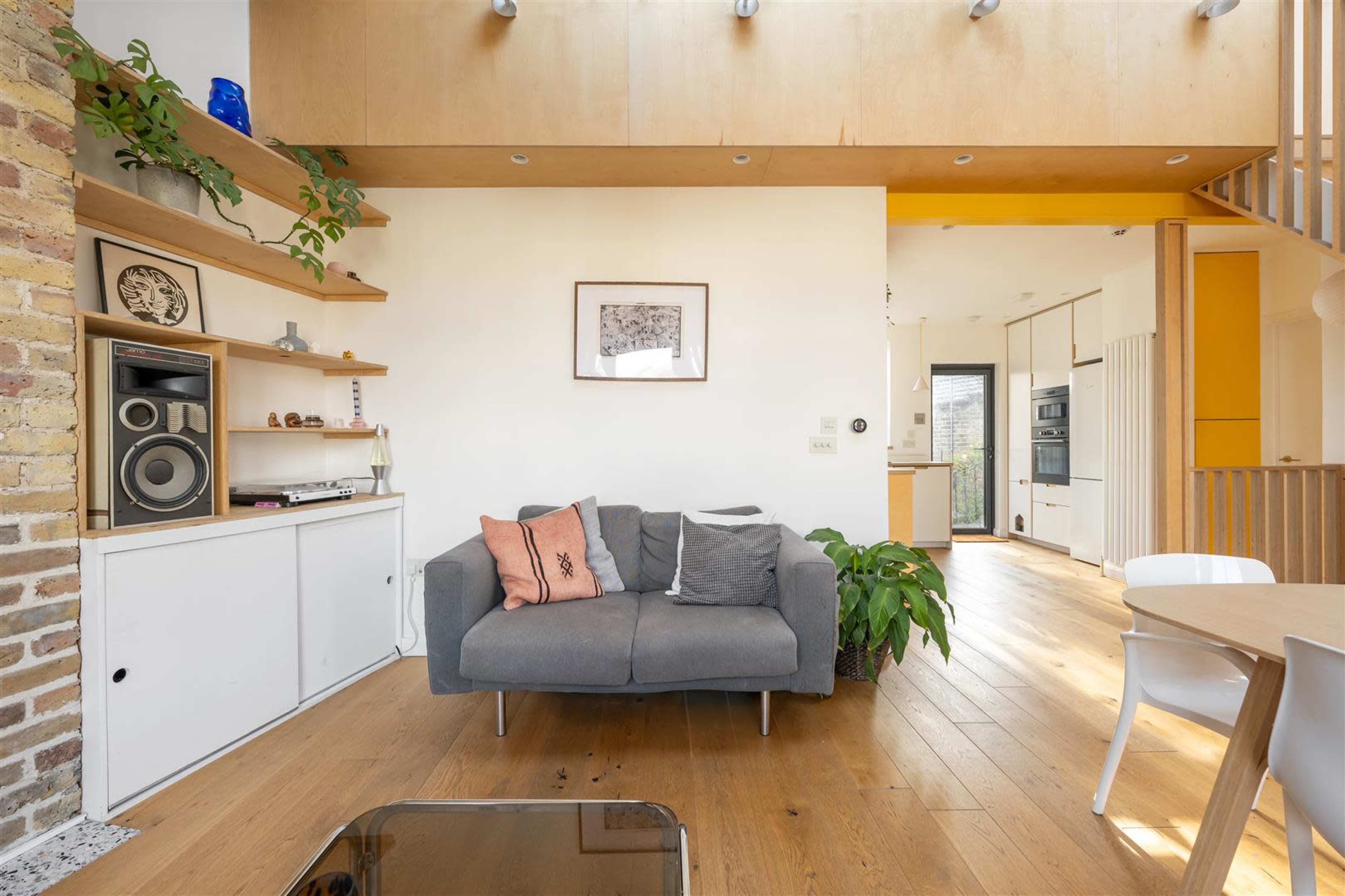 The image shows a modern living area with a gray sofa, wooden shelving, and a dining table, featuring large windows that allow natural light into the space.