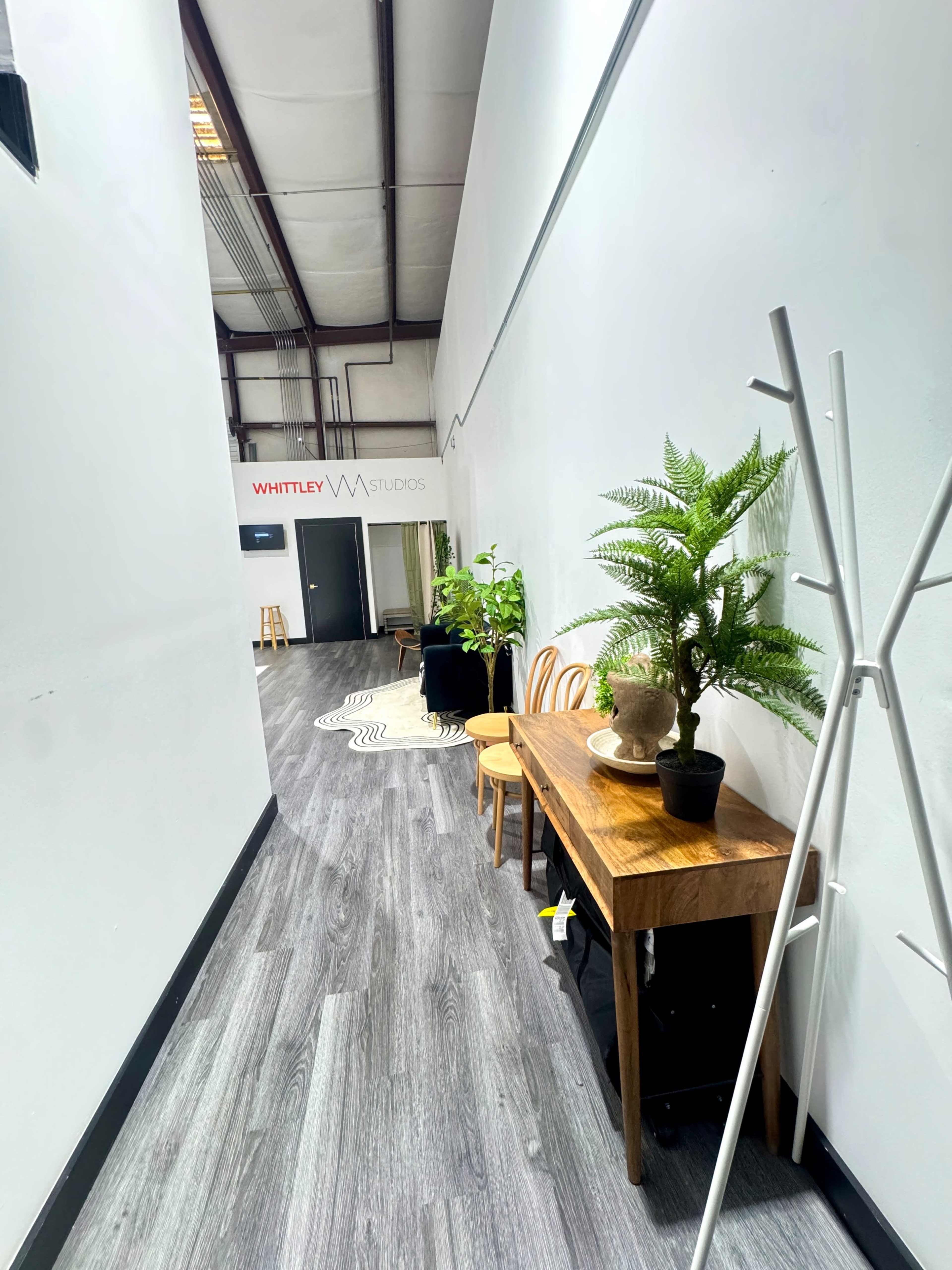 The image shows a long, narrow hallway with a wooden table, chairs, plants, and a door at the end labeled "WHITLEY Studios."