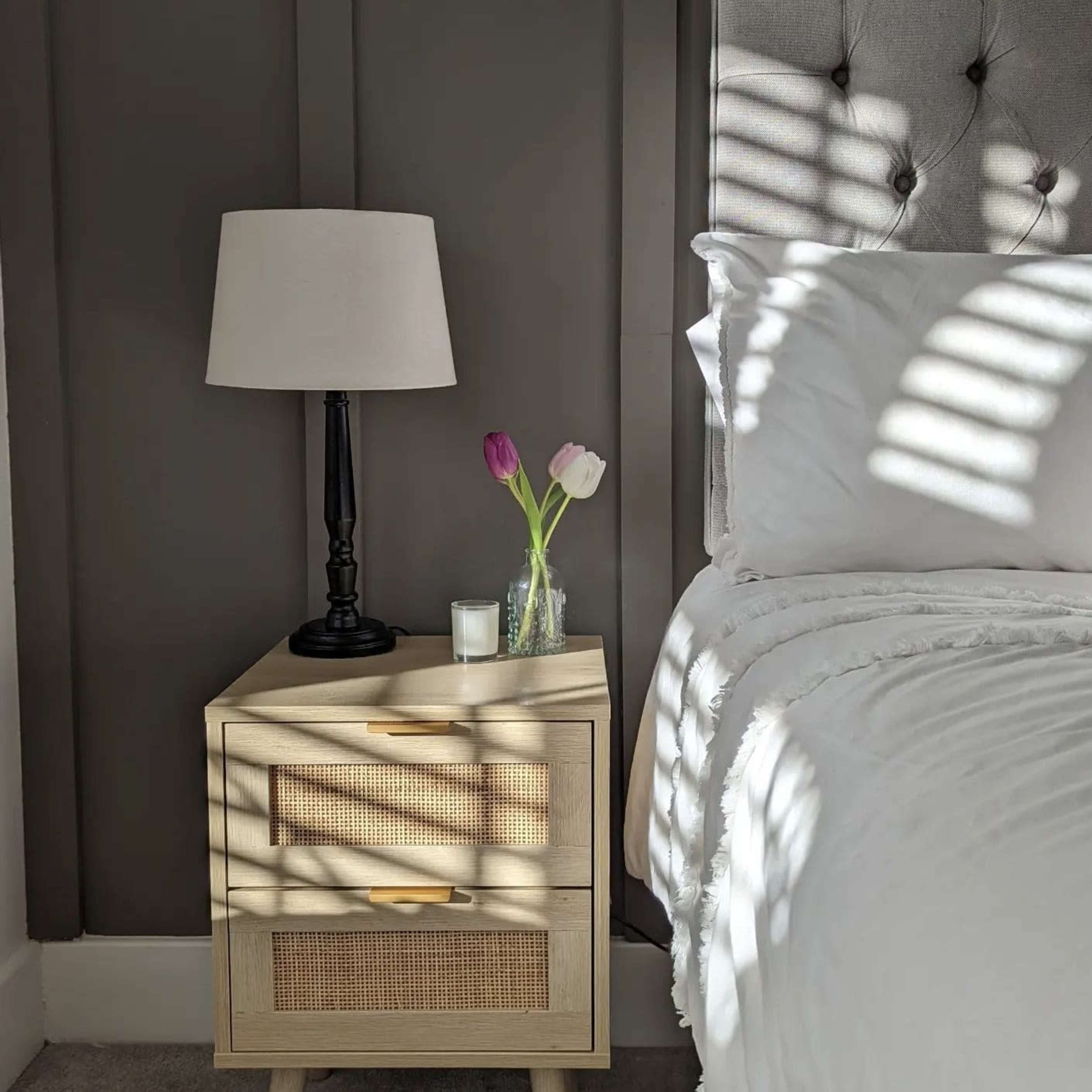 A bedside table with drawers, a lamp, and a vase of tulips sits next to a neatly made bed.