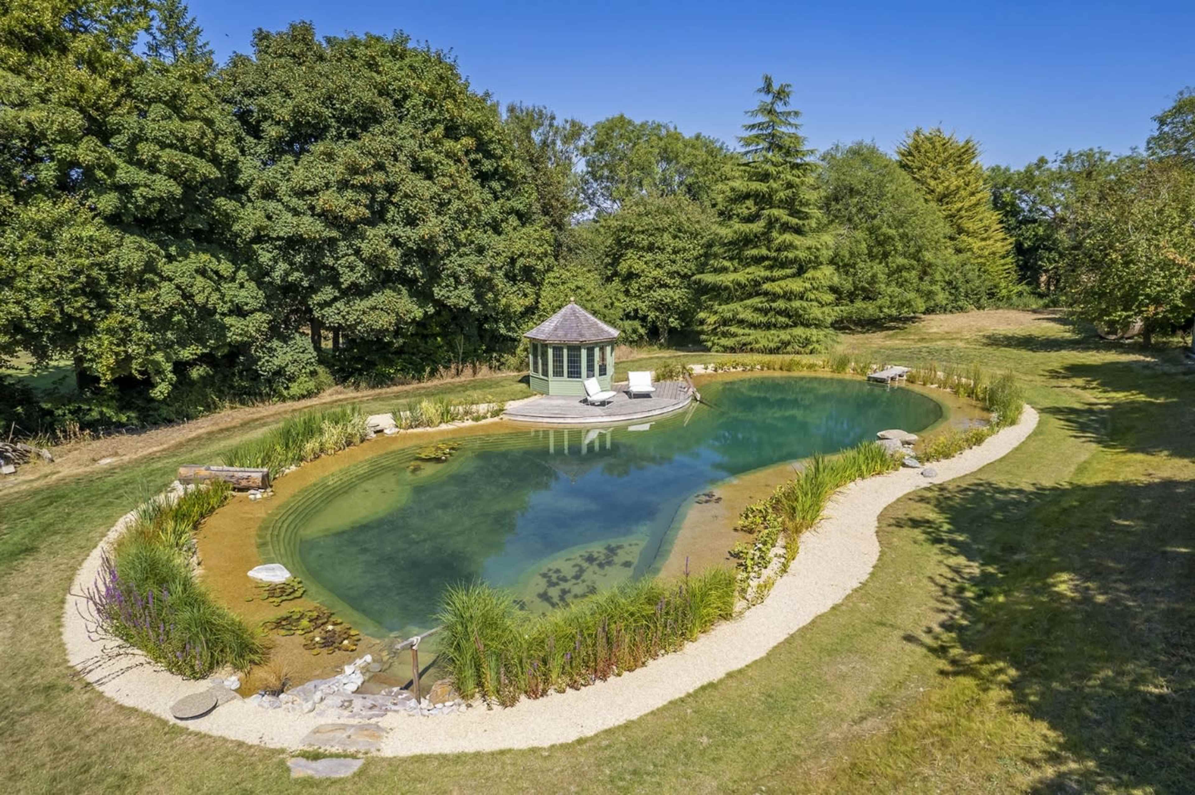 A peaceful pond is surrounded by lush greenery and features a circular patio with a gazebo at its center.