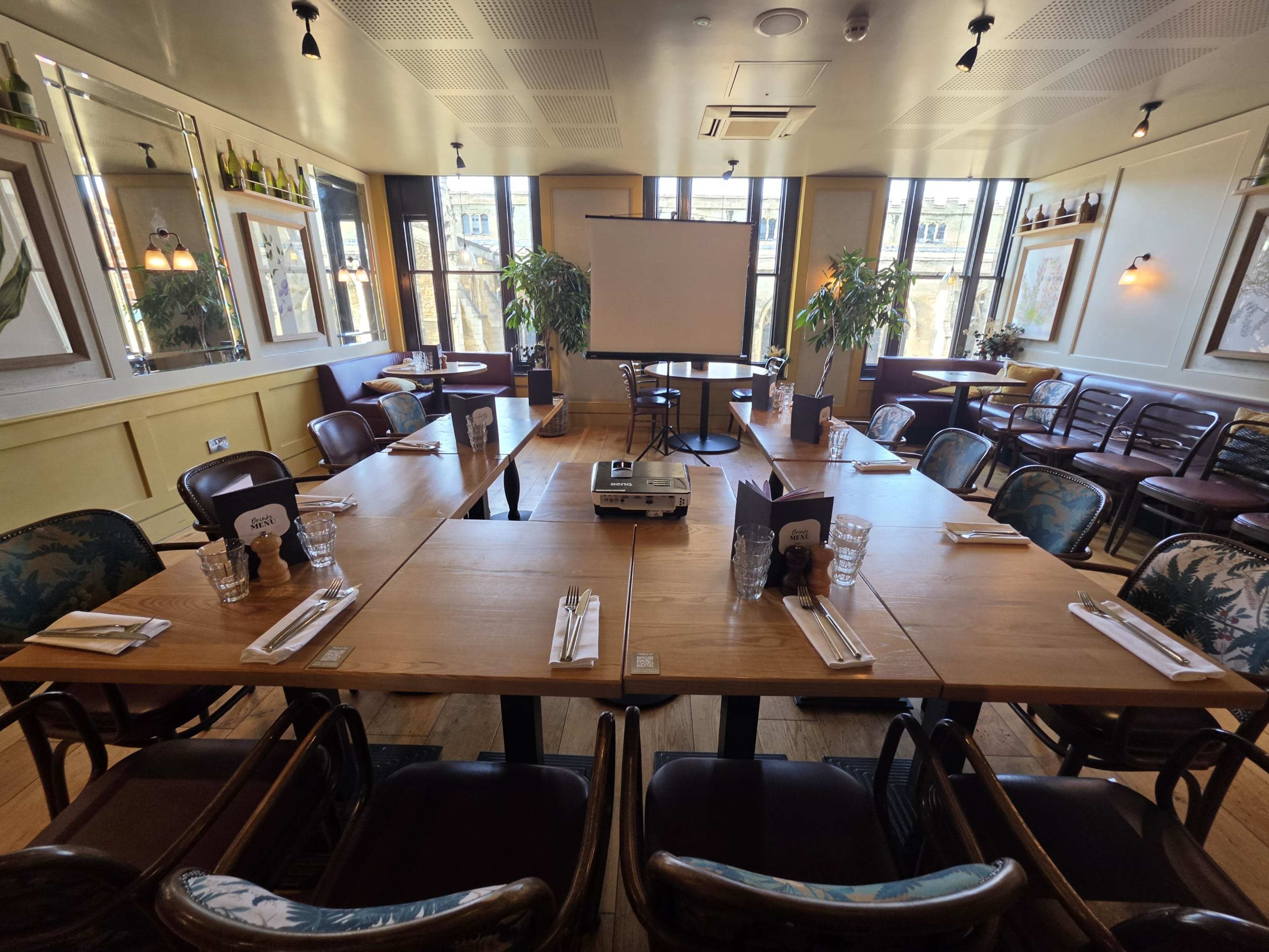 A brightly lit restaurant room features several tables arranged in a U-shape, with a projector screen positioned at the front.