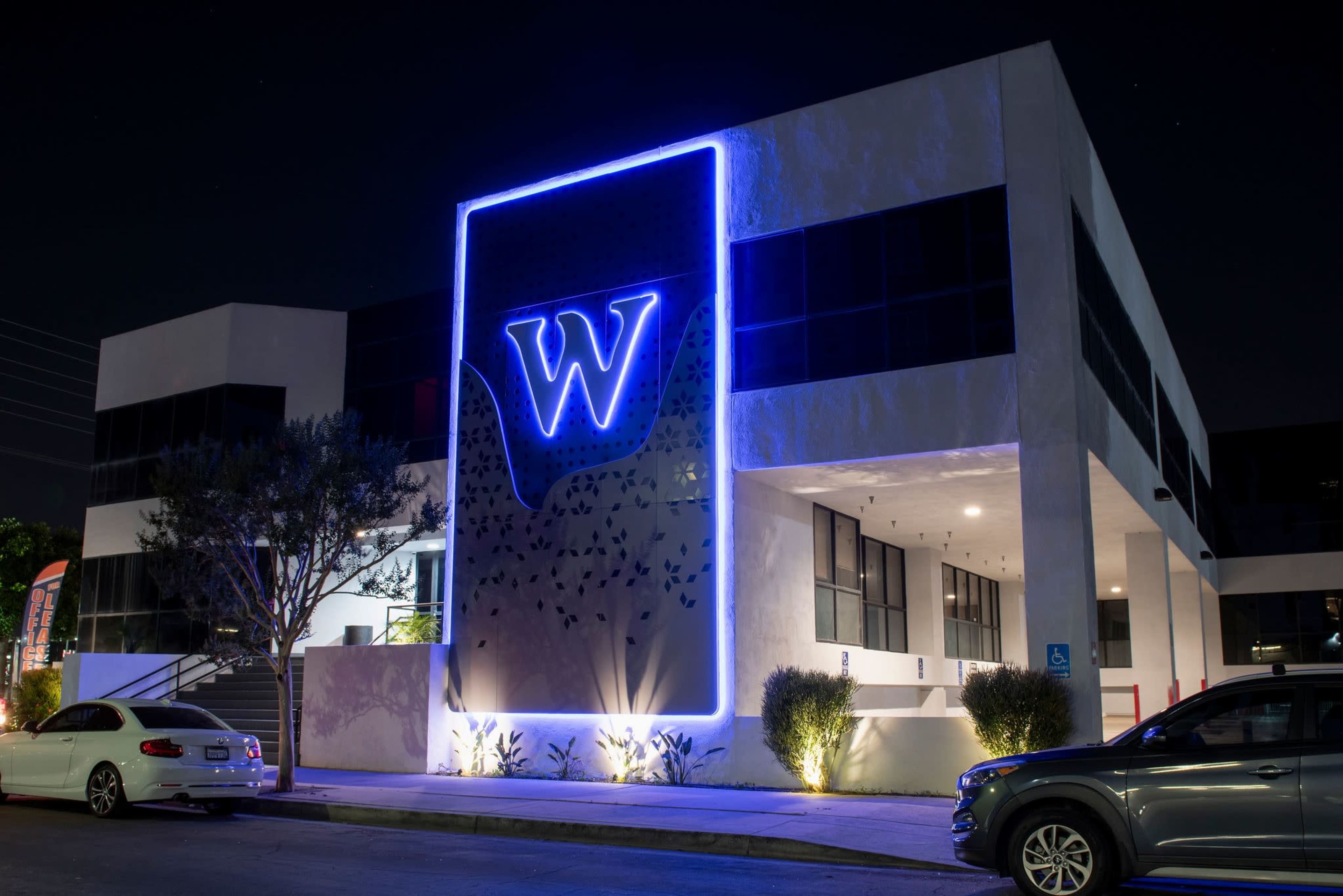 A modern building features a prominent illuminated "W" logo on its facade, surrounded by landscaped greenery at night.