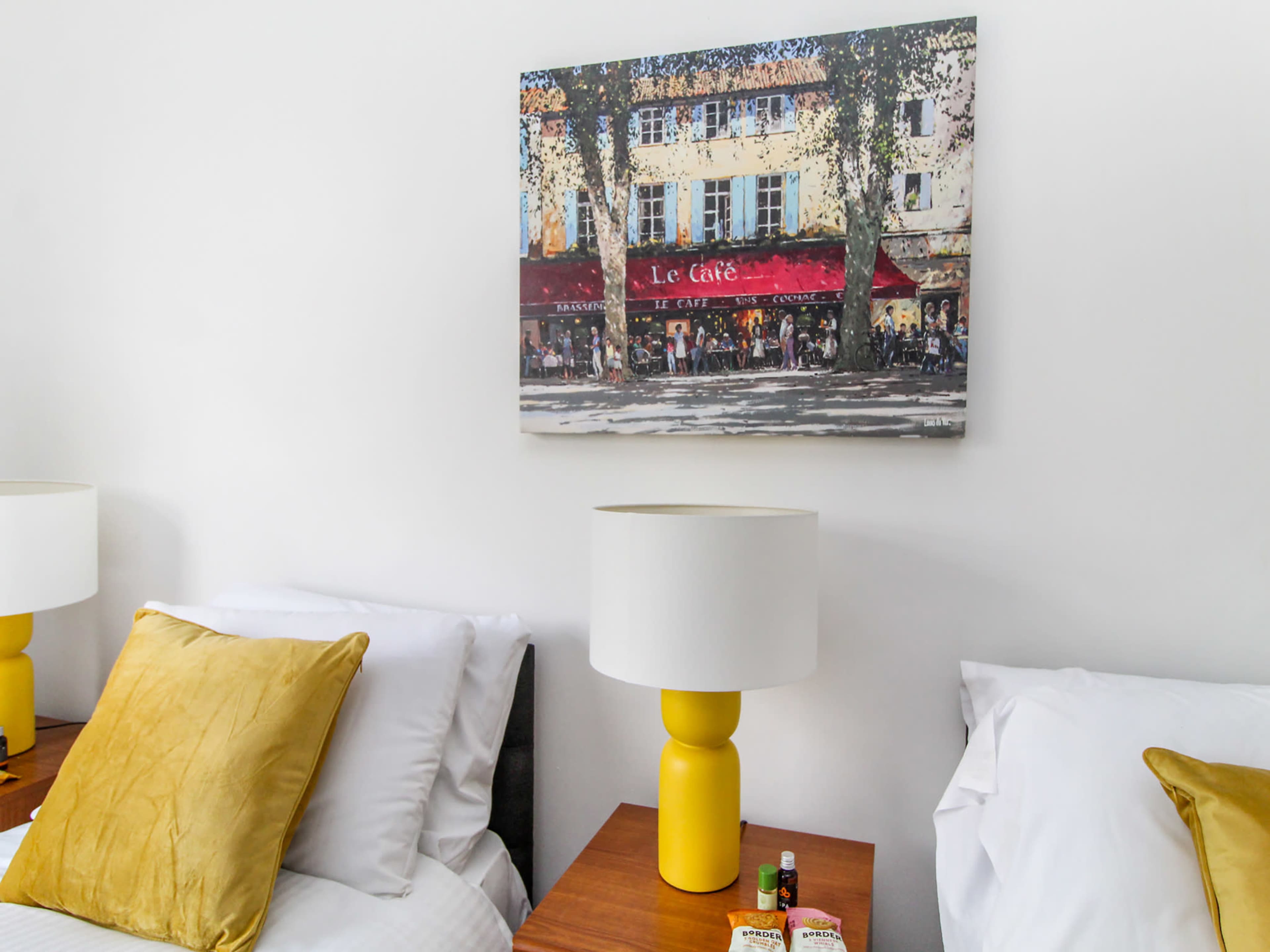 The image features a hotel room with two beds, each accompanied by yellow lamps, and a painting of a café on the wall above.