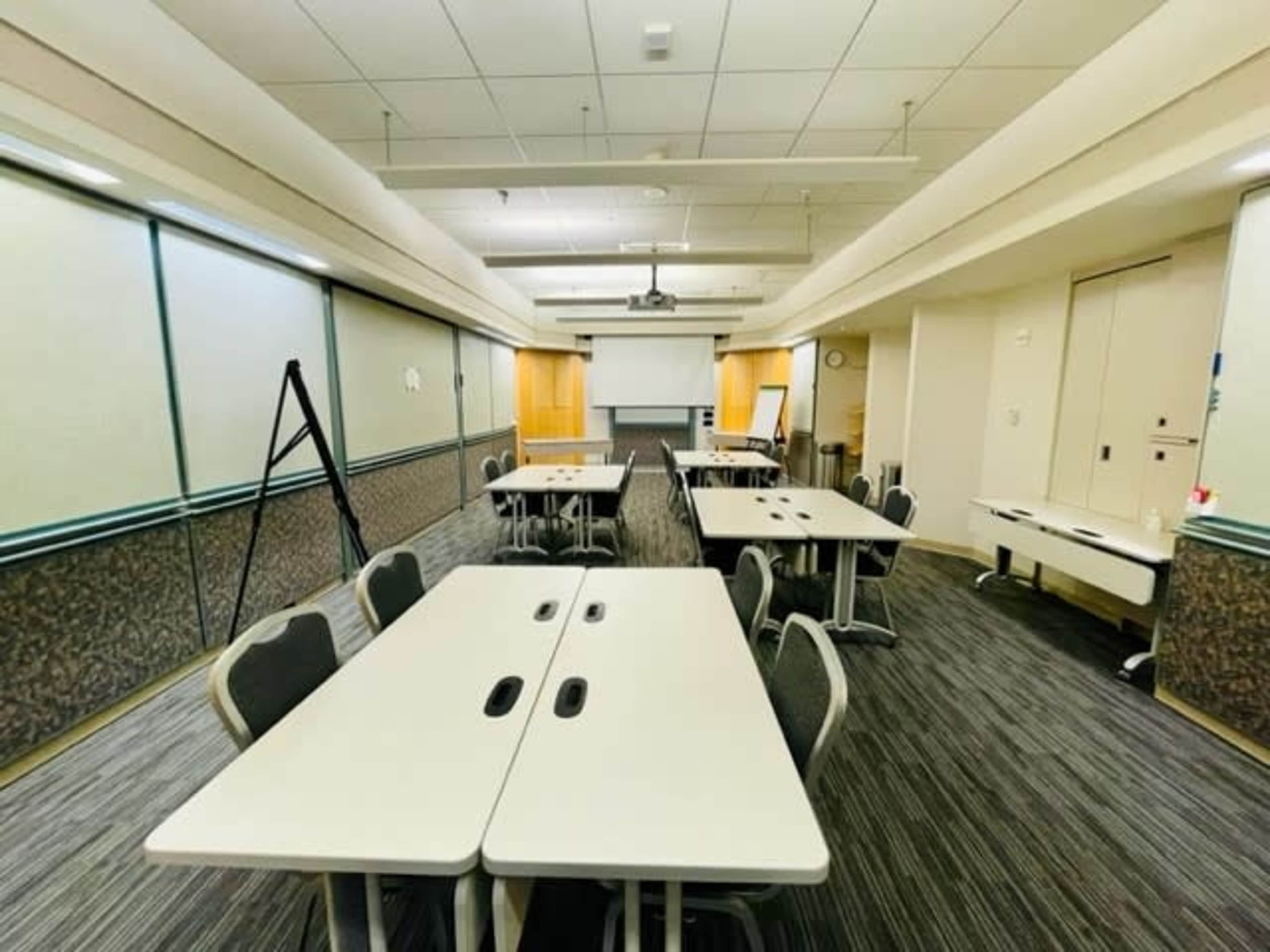 A spacious meeting room features several white tables arranged with chairs, soft lighting, and a projector screen at the front.