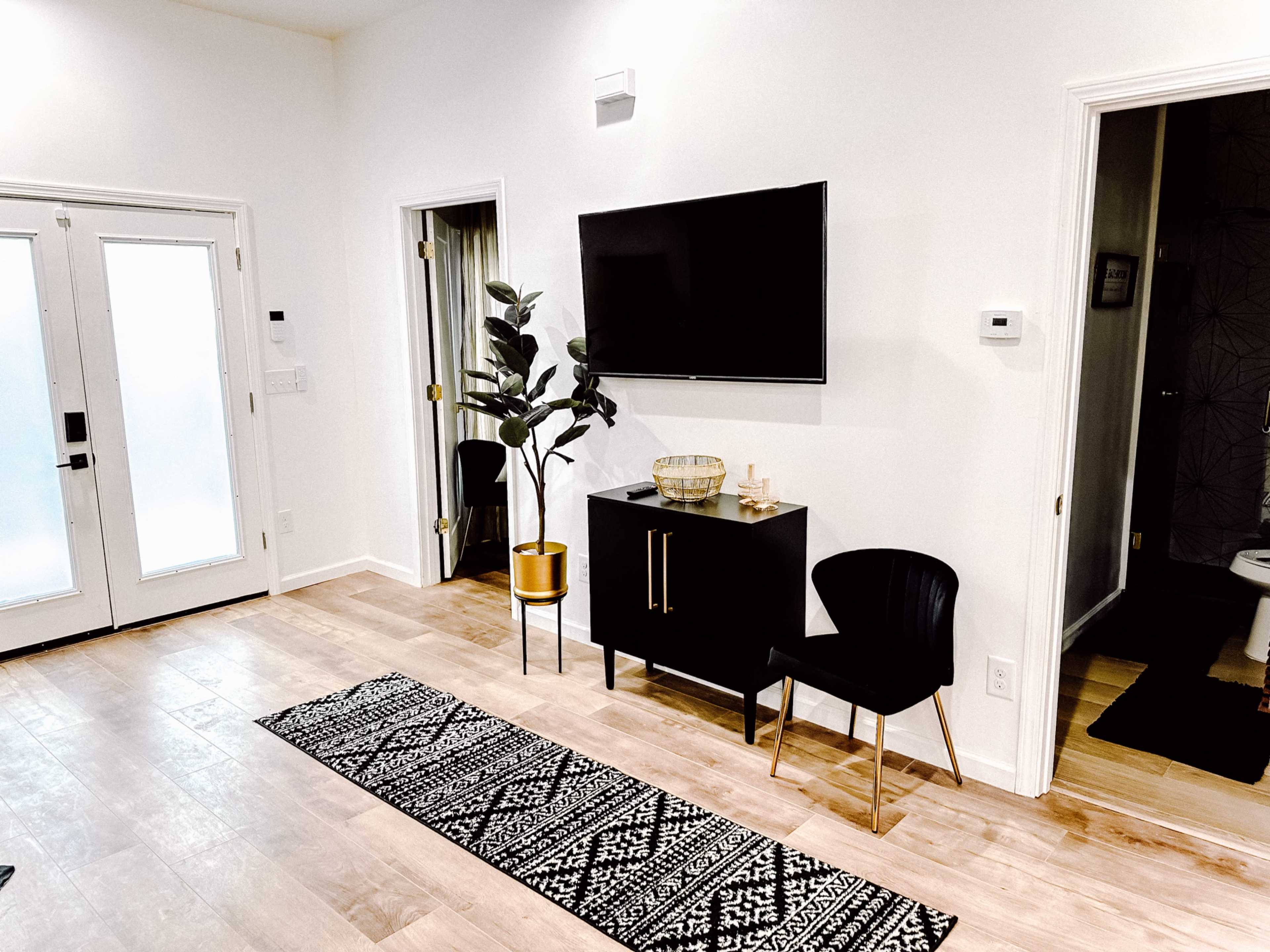 The image depicts a modern living room featuring a wall-mounted television, a black cabinet, a chair, a patterned runner rug, and a plant next to a set of double doors.