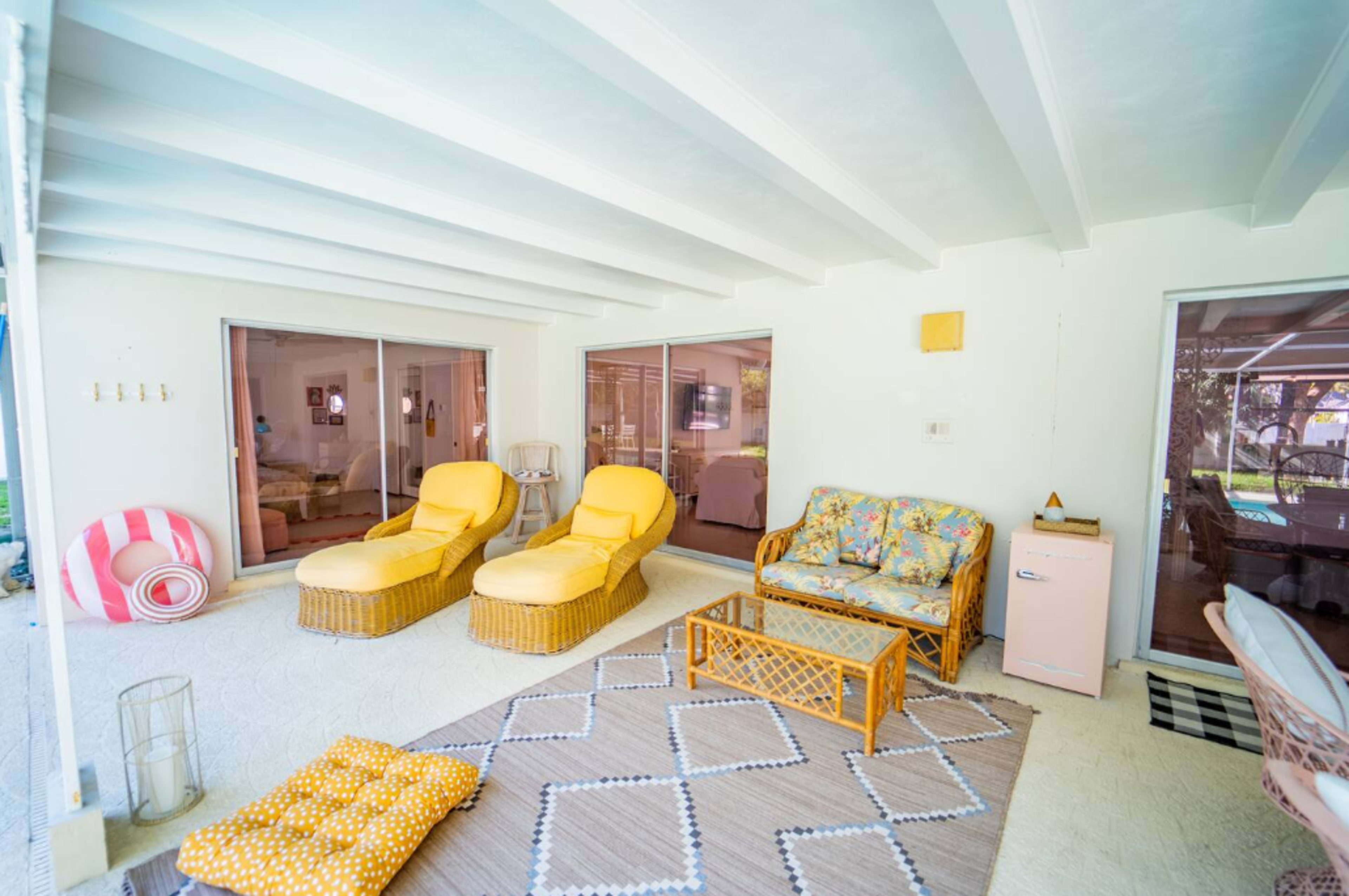 The image shows a bright sunroom with two yellow lounge chairs, a patterned loveseat, and a braided rug on the floor.