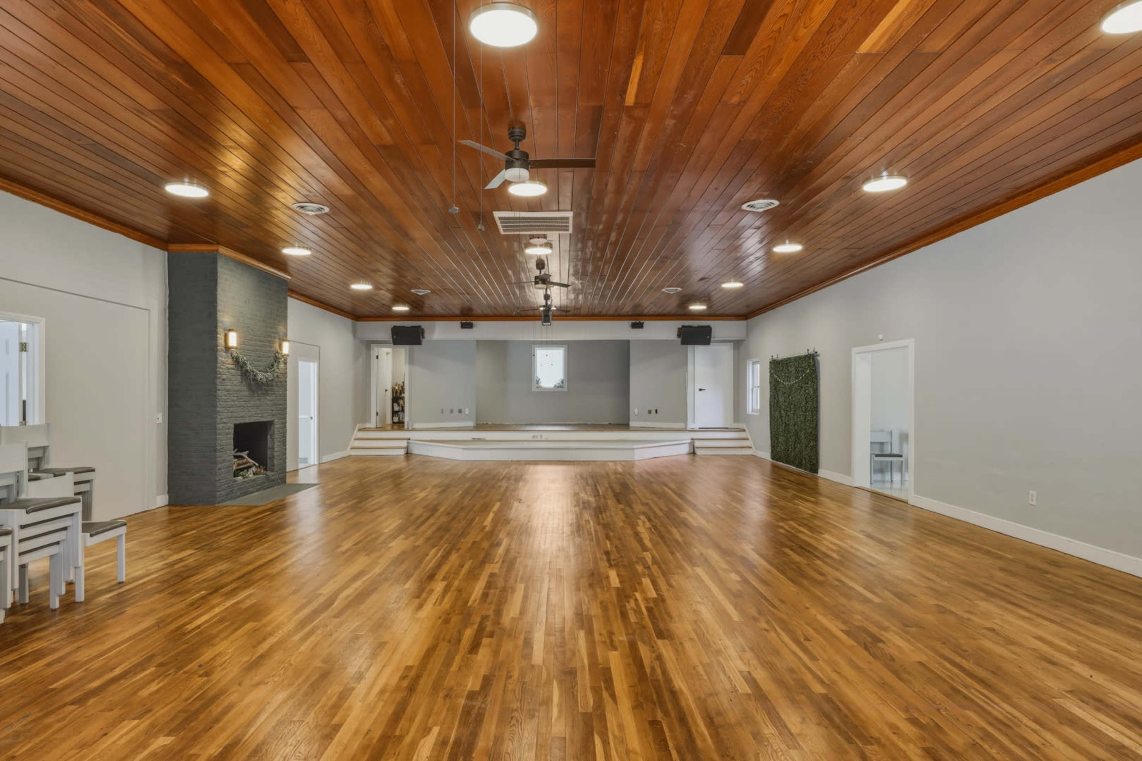 The image shows a spacious interior room with a wooden ceiling, hardwood flooring, and a raised stage at one end.