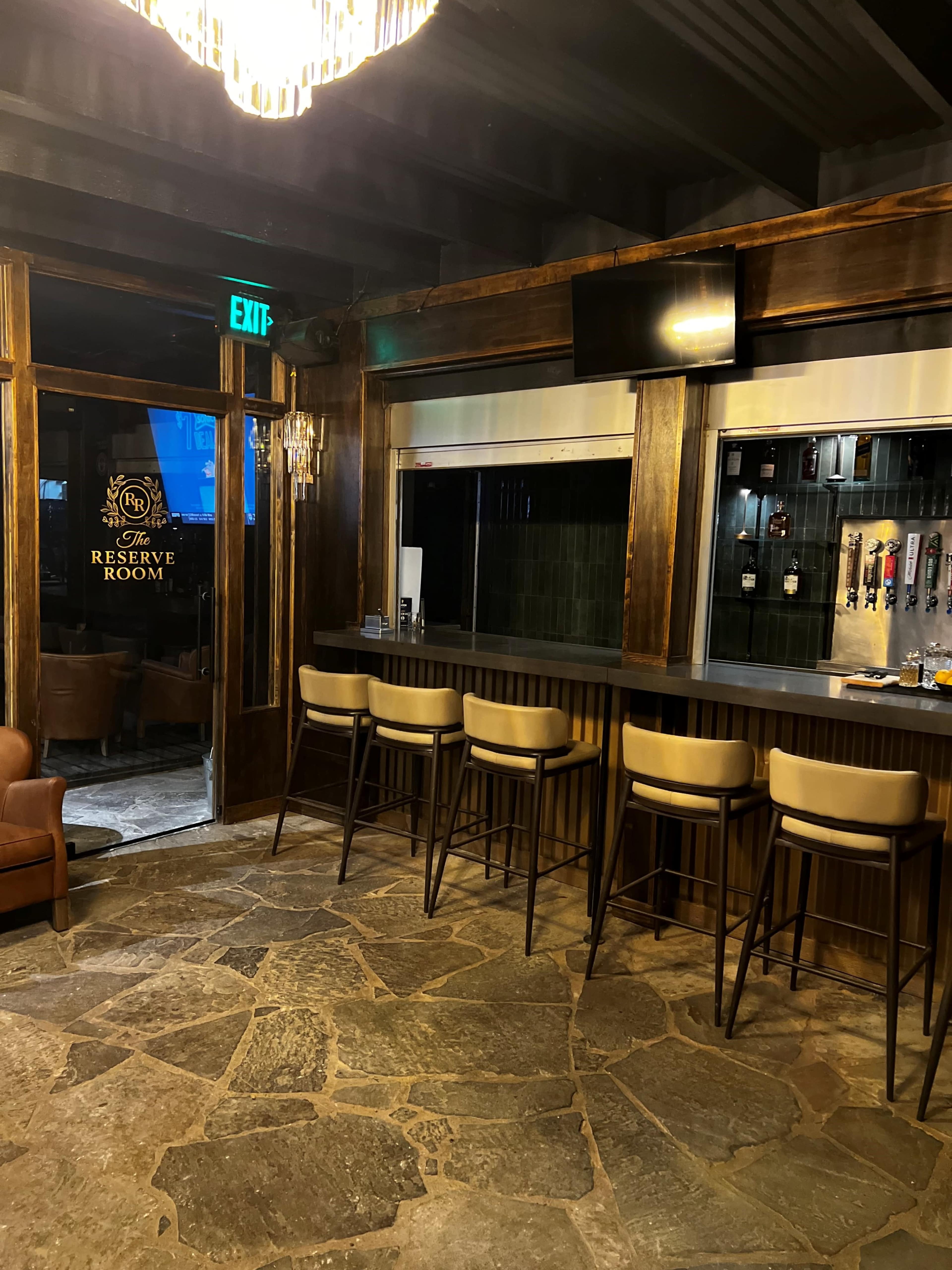 The image shows a dimly lit bar area with stone flooring, wooden paneling, and high stools arranged in front of a counter that features tap beer faucets and a television screen.