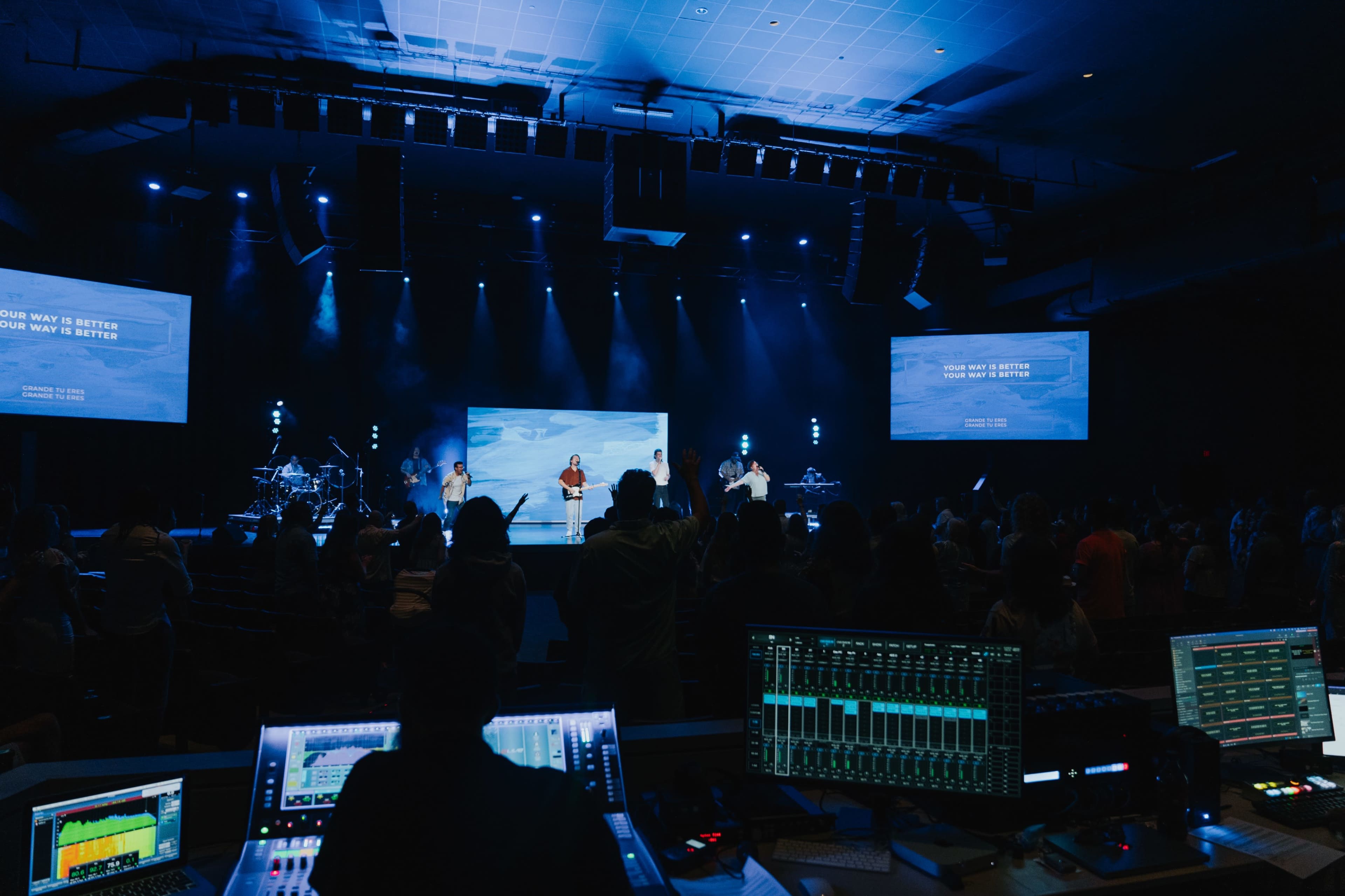 A group of performers are on stage in a lit auditorium with a live band setup, while an audience participates and a soundboard is visible in the foreground.