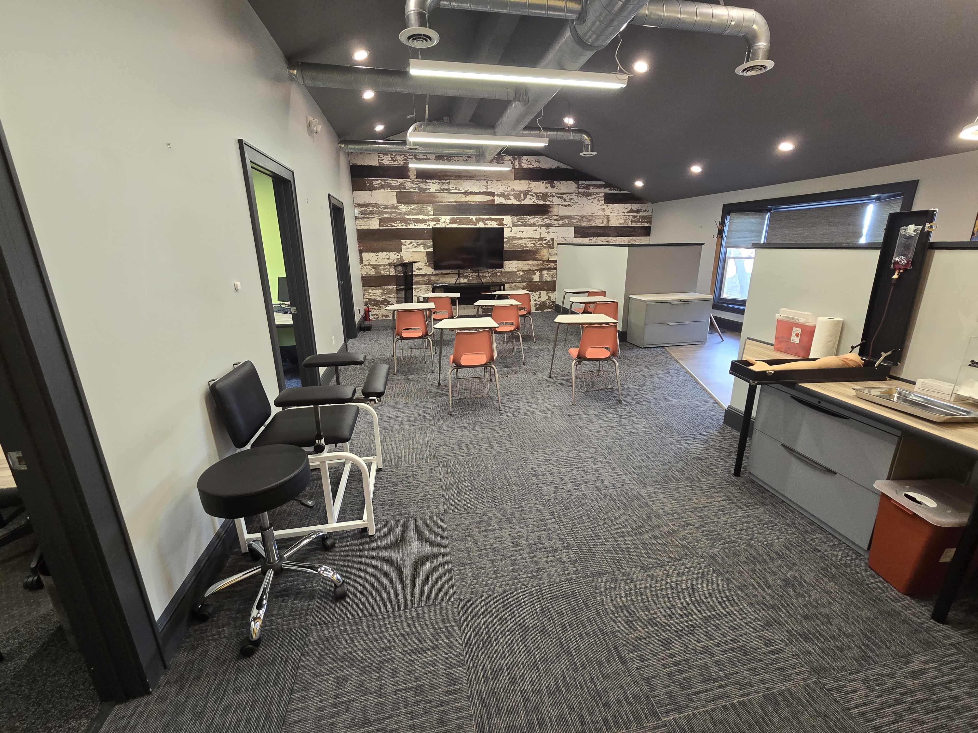The image shows an office space featuring gray carpeting, a rustic wood accent wall, several tables with chairs, and a television mounted on the wall.