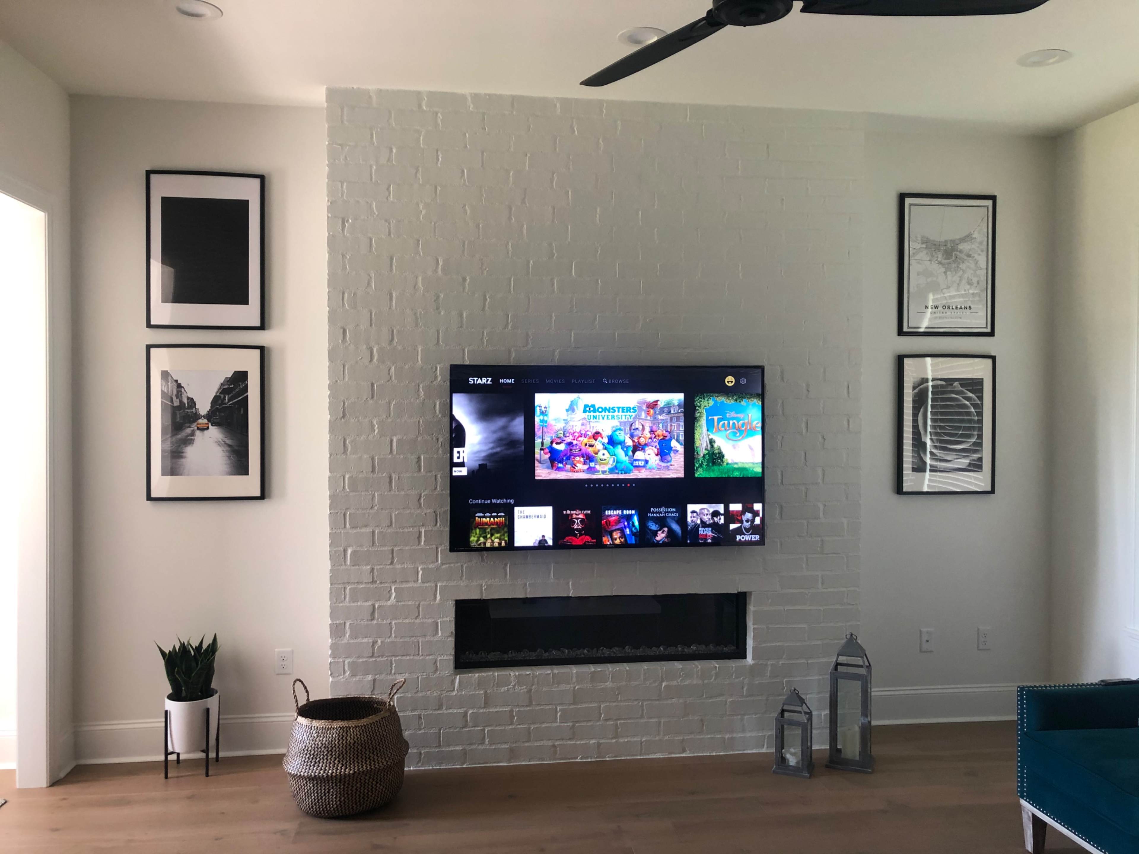 A living room with a white brick wall featuring a mounted television, framed artwork on either side, and a decorative arrangement including a plant and lanterns below the TV.