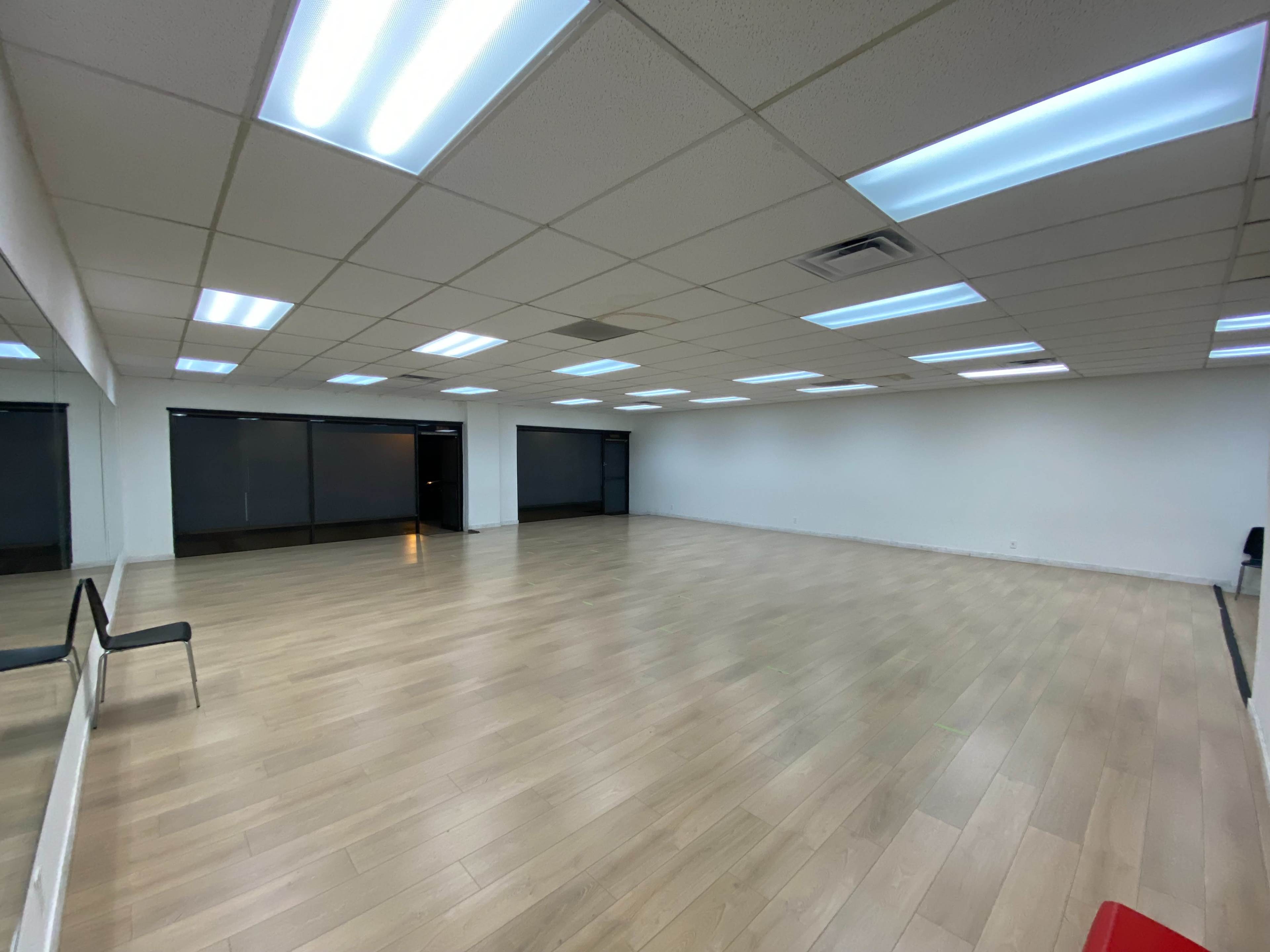 A spacious interior room with wooden flooring, fluorescent ceiling lights, and large windows along one wall.