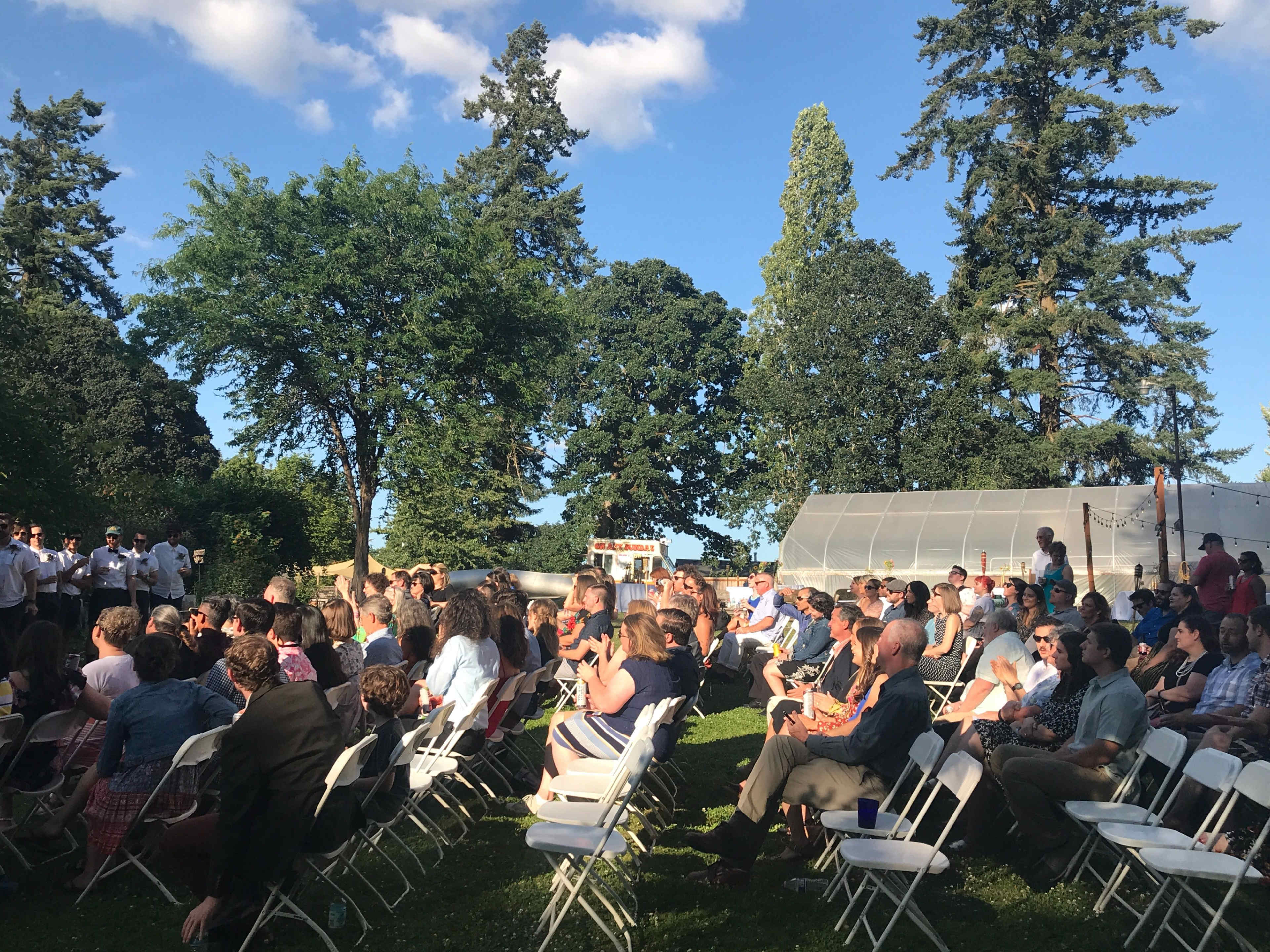 A large crowd of people sits in white folding chairs under trees during an outdoor event.