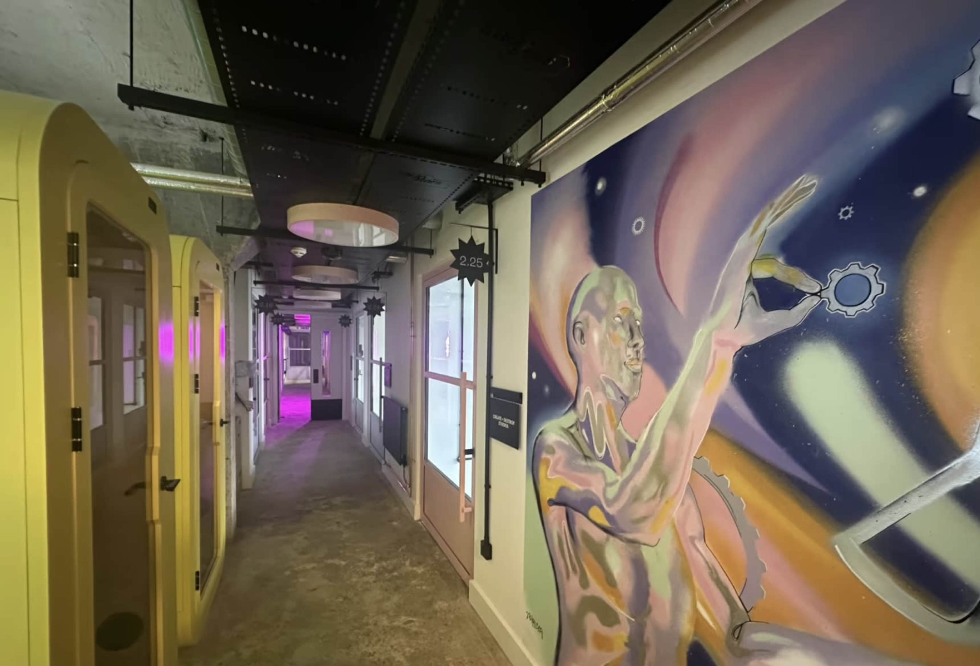 The image shows a modern, colorful hallway featuring a mural of a figure reaching toward an abstract background, with yellow phone booths on one side.