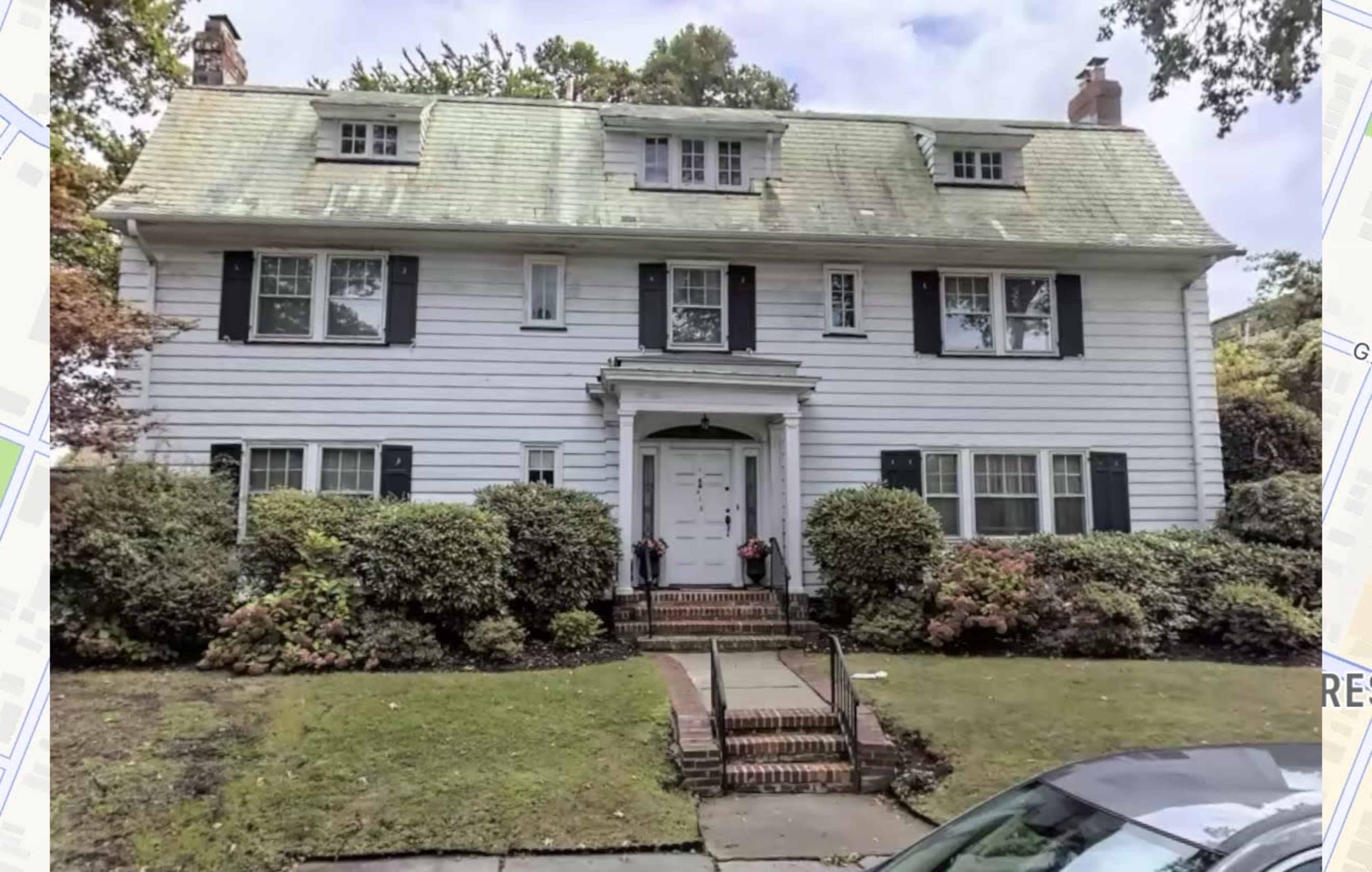 The image shows a large two-story house with white siding, a steeply sloped roof, and a brick walkway leading to the front door, surrounded by shrubs and greenery.