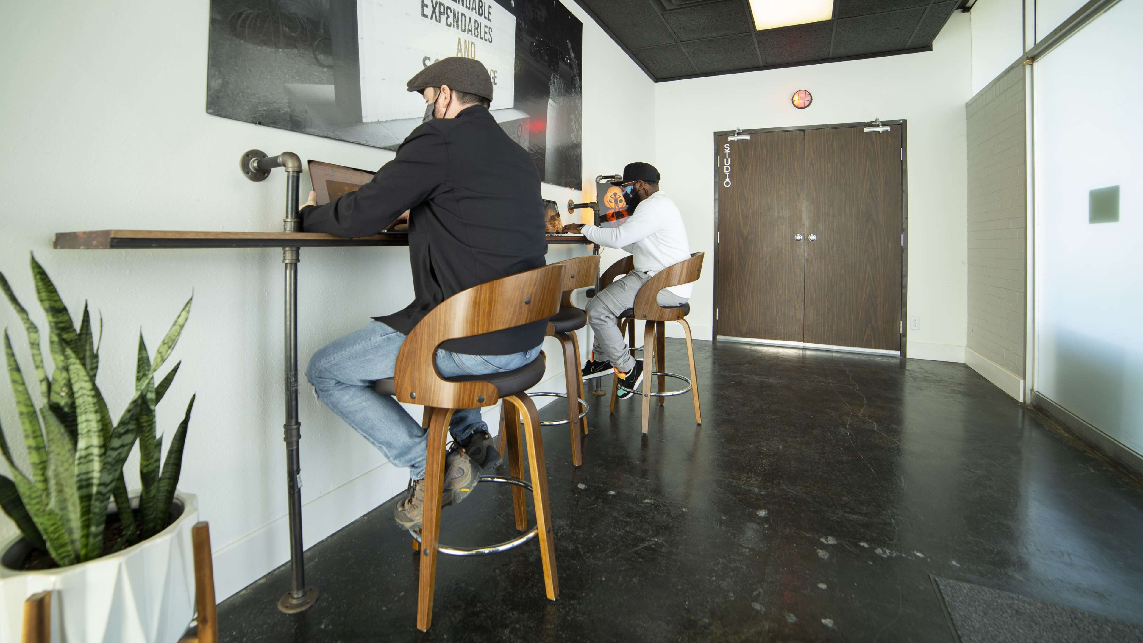 Two men sit on high stools at a minimalistic workspace, focused on their laptops against a white wall.
