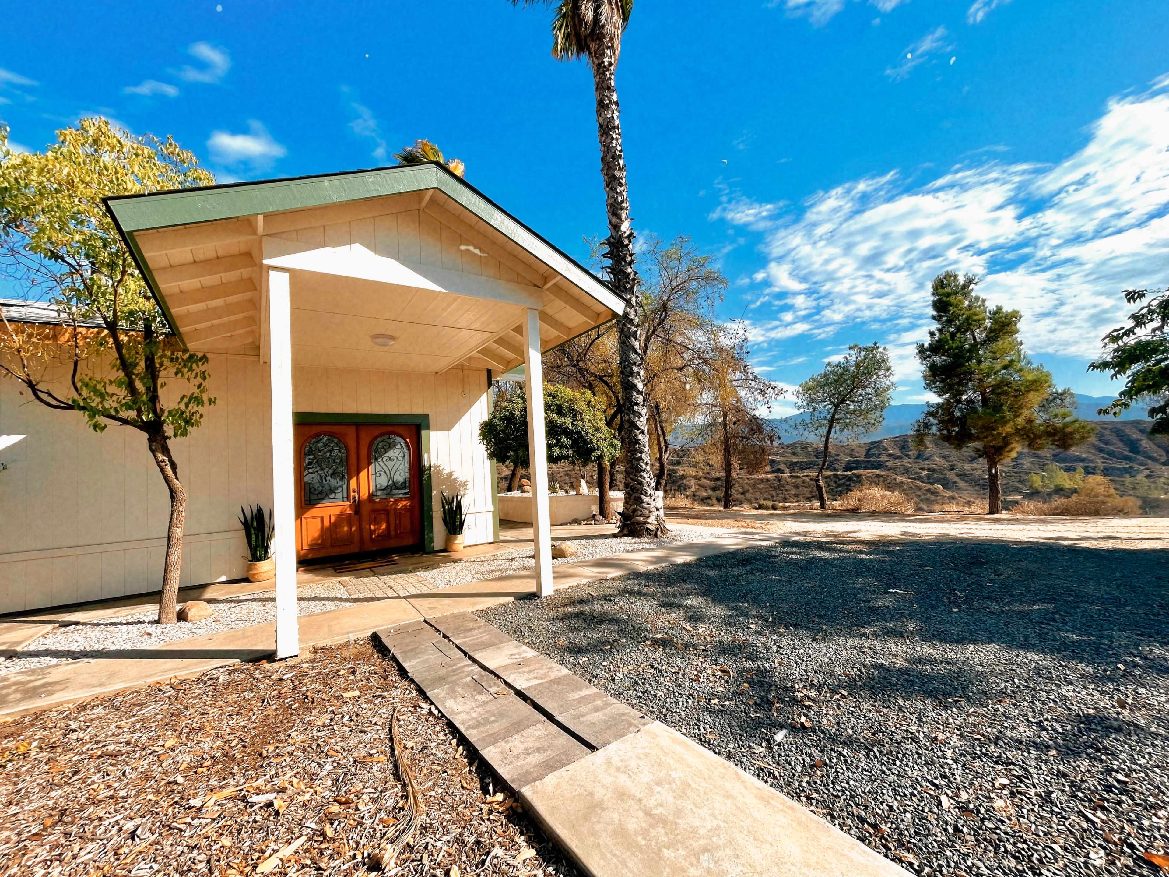 A modern house with a wooden double front door sits under a clear blue sky, surrounded by gravel and sparse trees.