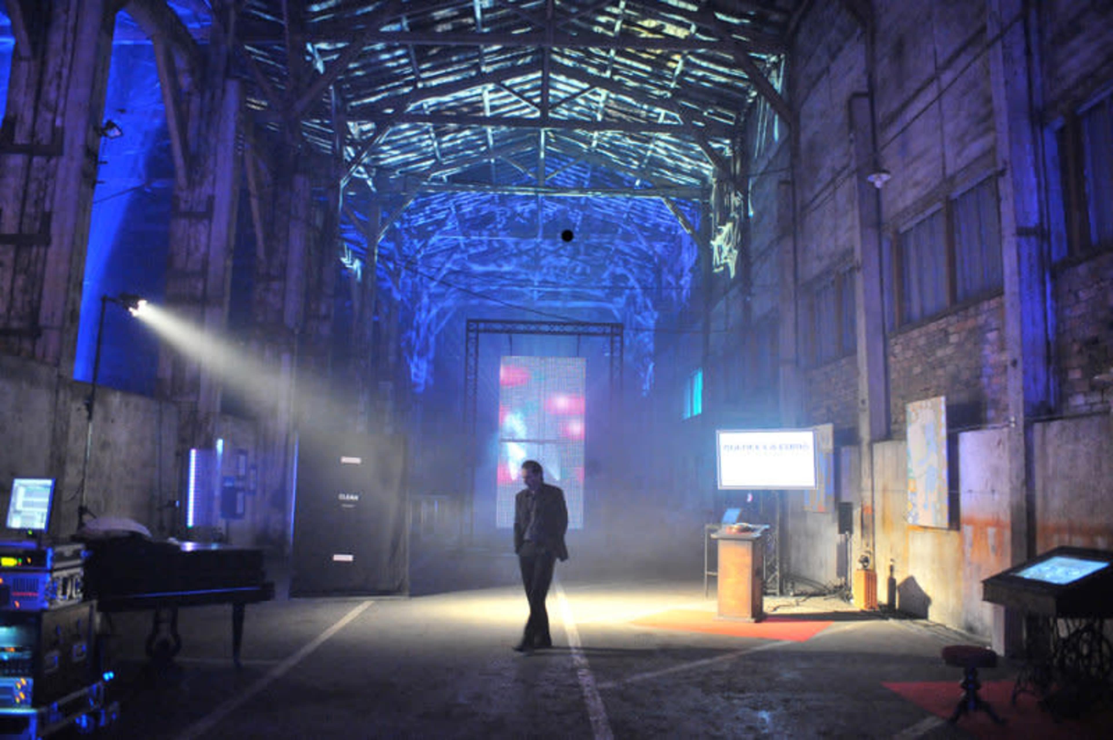 The image shows a spacious, industrial warehouse with blue lighting, fog, and a person standing in the center.