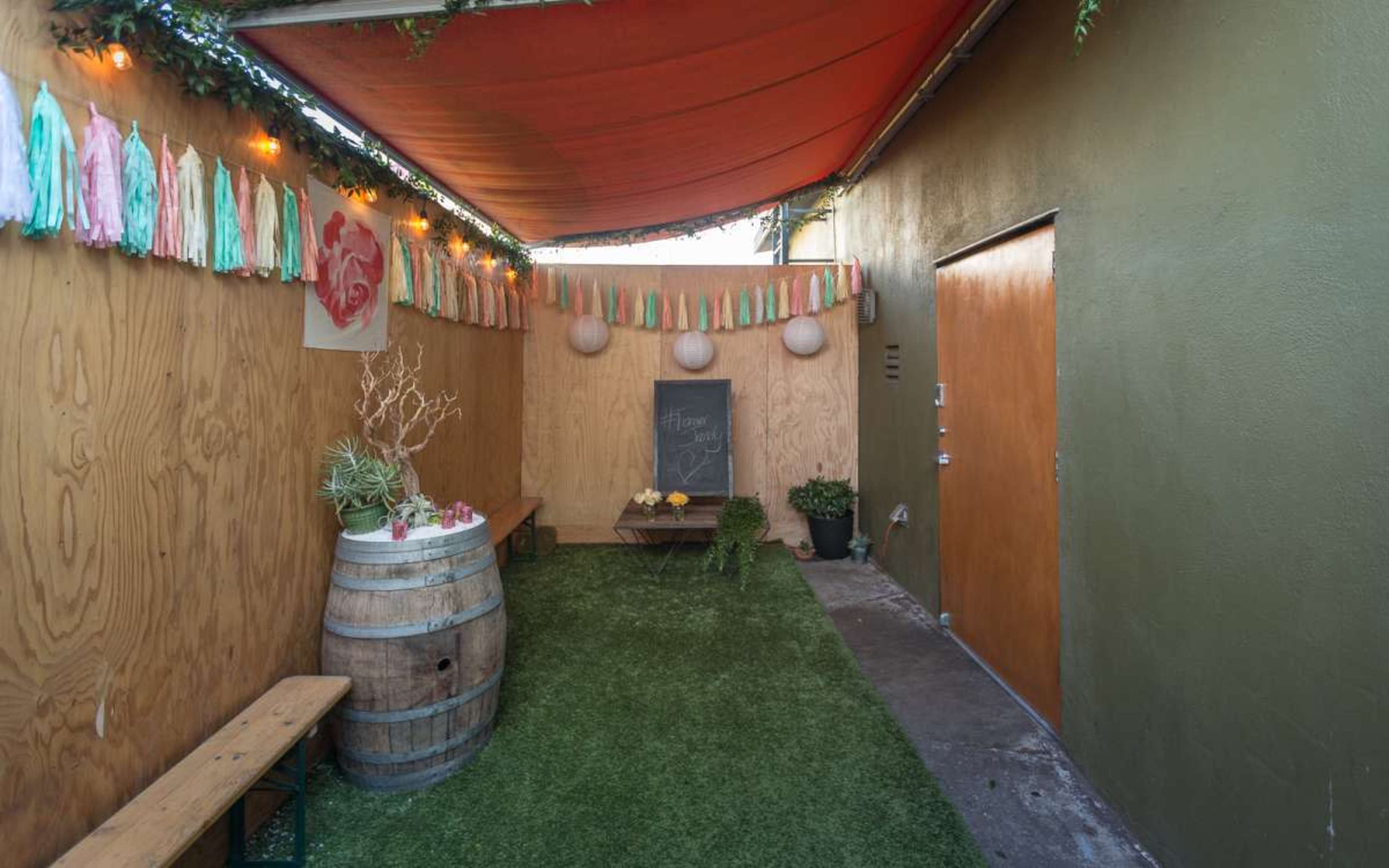 The image shows a narrow outdoor space with a wooden wall, a barrel table, a bench, and decorative streamers hanging from the ceiling.