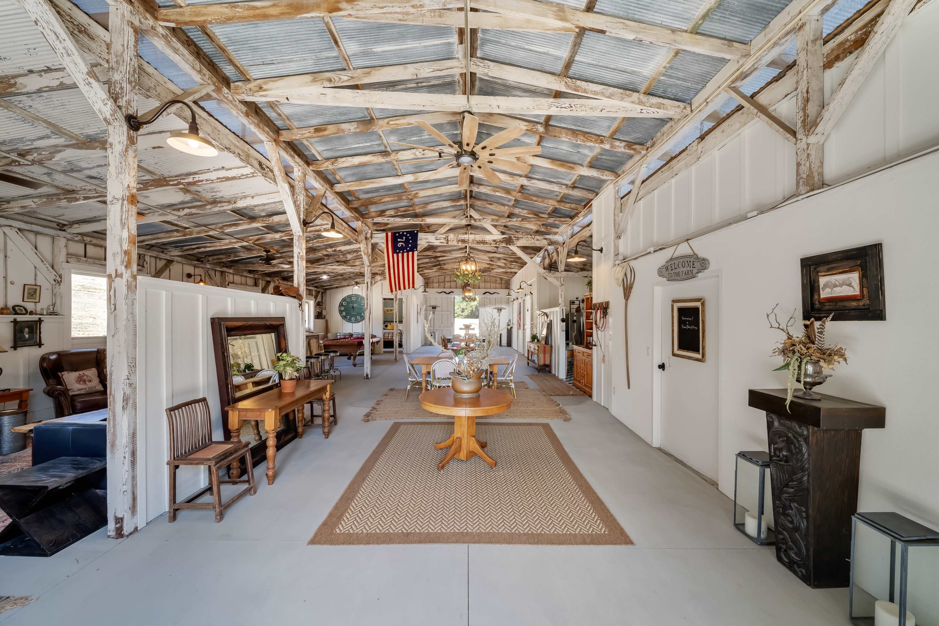 The image depicts a spacious indoor area of a rustic building with exposed wooden beams, an American flag hanging from the ceiling, and various furniture pieces arranged along the sides.